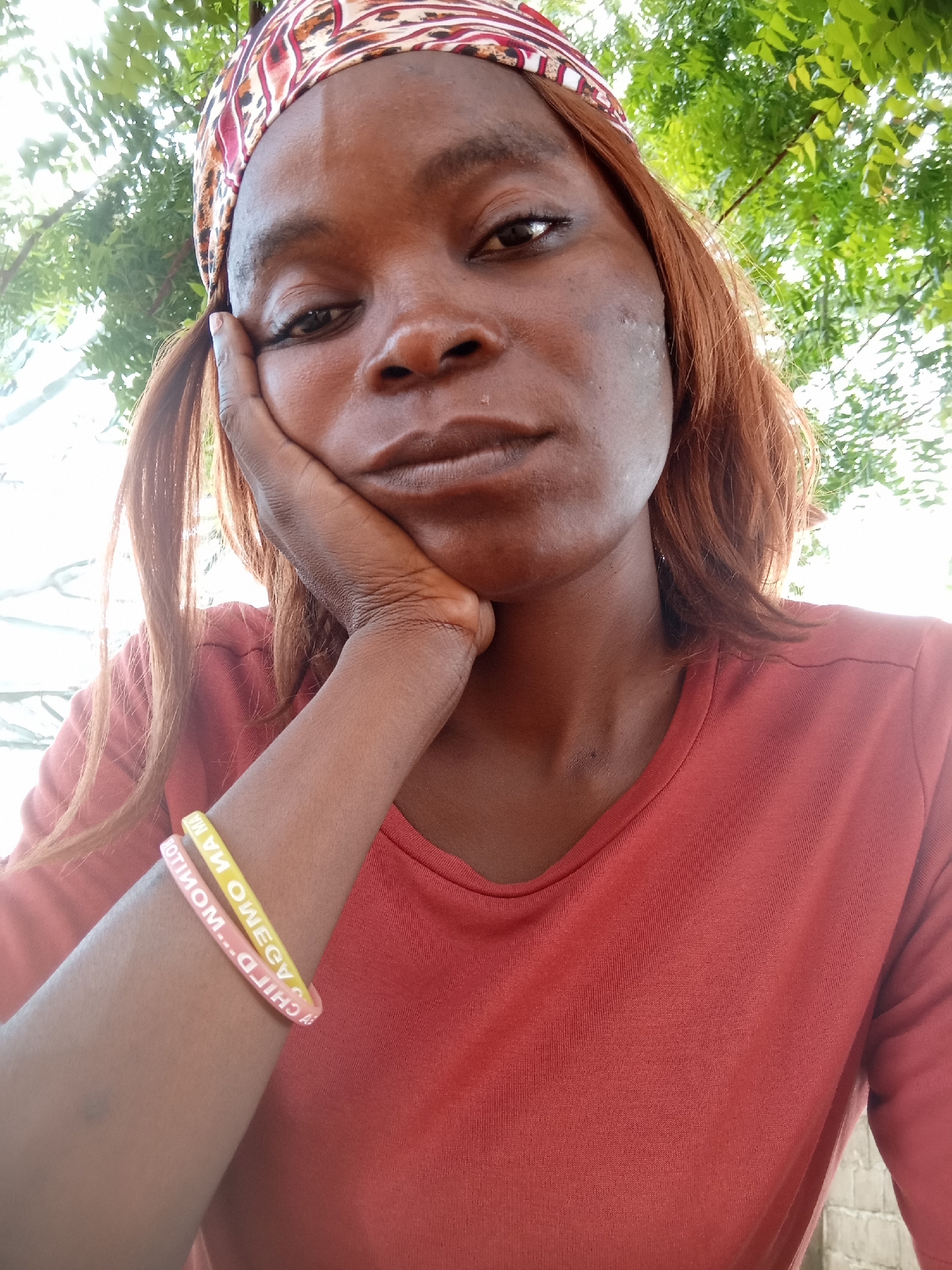 A medium shot shows a young Black woman from the chest up, with her head tilted slightly to her left and her right hand propping up her chin. She is wearing an orange crew-neck t-shirt and a colorful bandana on her head. Her hair is dark brown and falls to her shoulders. She is wearing two rubber bracelets on her left forearm, one pink and one yellow, with white lettering on them that reads "CHILDREN MATTERS" on the pink one and "WOMEN MATTERS" on the yellow one. The background consists of green foliage and bright sky, suggesting an outdoor setting. The woman appears to be in Kason, Nigeria, as the image was geolocated there.