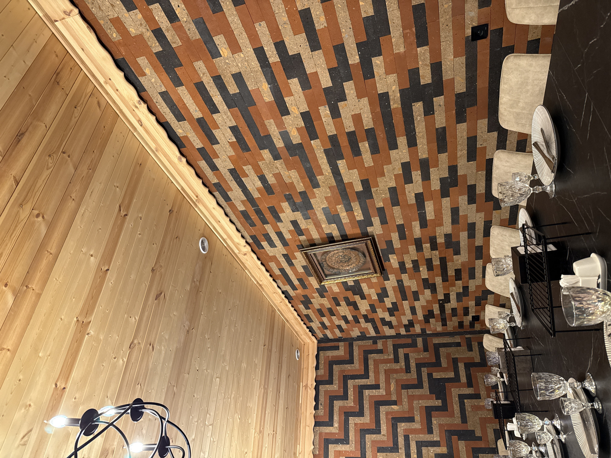 The image shows an interior space with a combination of wood paneling and patterned brickwork. The left wall is covered in vertical wood panels. Above, the ceiling features a pattern of alternating black and terracotta bricks in a running bond arrangement, with a framed decorative artwork centered on the ceiling. The floor is paved with the same bricks, laid in a zigzag pattern. Along the right side, a dark marble table is set for a meal with chairs, plates, napkins, water glasses and serving stands. A modern chandelier with lights is partly visible in the lower left. The location is Arinj, Armenia.