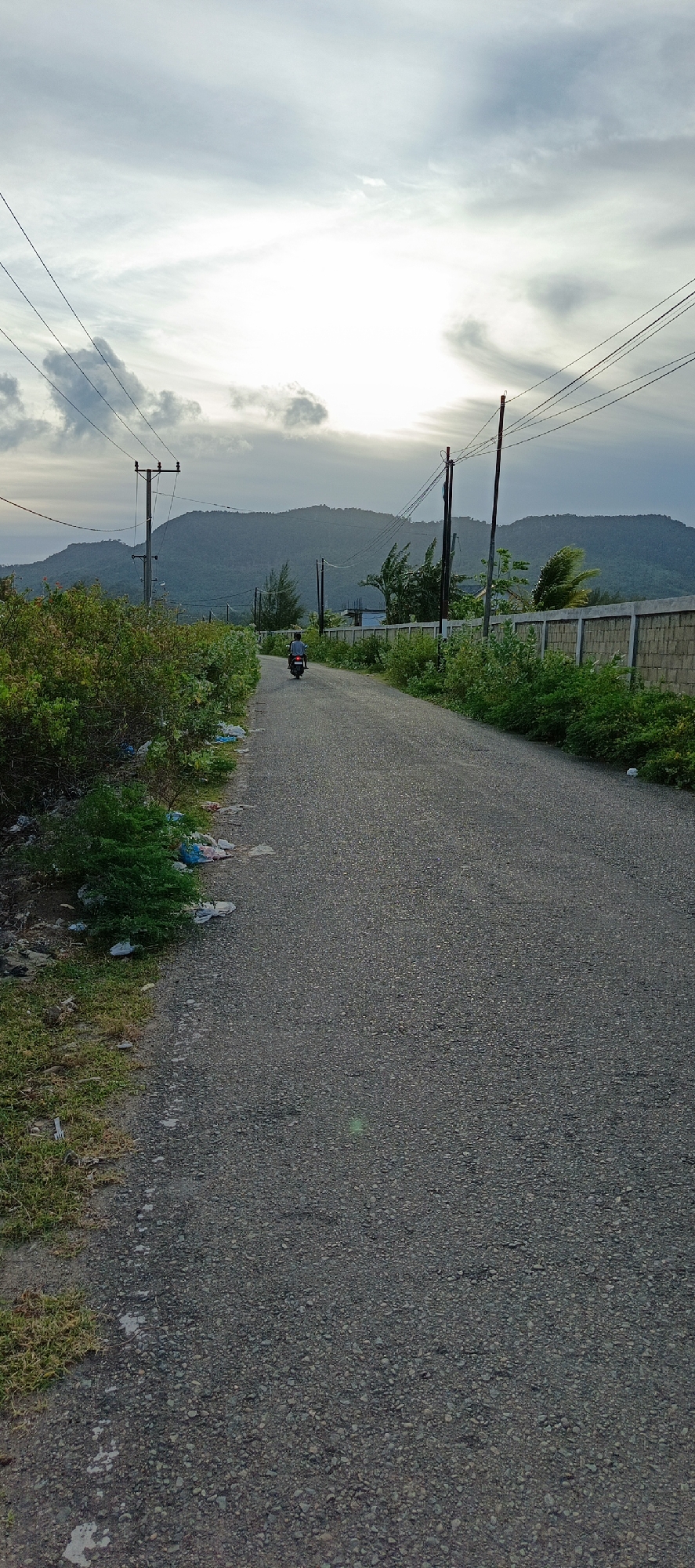 The image depicts a paved road in Lamguron, Indonesia, under a cloudy sky. A lone individual is riding a motorcycle away from the viewer down the road. Lush green vegetation lines the left side of the road, with some scattered trash visible. On the right side, a stone wall borders the road, with more greenery behind it. In the distance, a large, verdant hill or mountain range dominates the landscape, partially obscured by the hazy sky and the sun's glow. Electrical poles and wires are visible along the road, suggesting the presence of infrastructure. The overall scene conveys a rural or semi-urban setting, possibly during the late afternoon or early morning, given the lighting.
