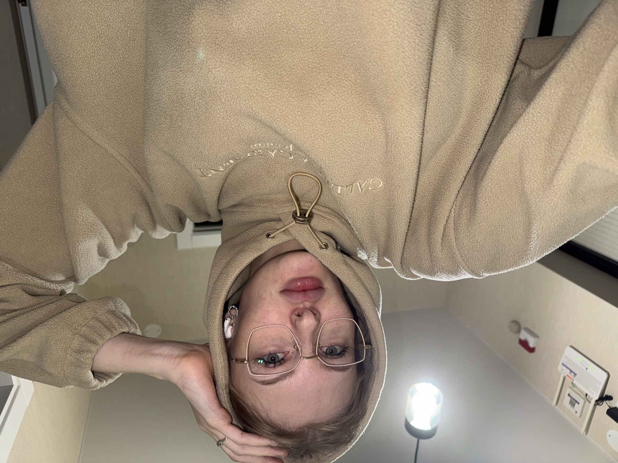 A person is upside down in the photo, wearing eyeglasses and a beige fleece hoodie with the word "GARMENT" partially visible. They have one hand near their head. An earbud is visible in their left ear. The room appears to be indoors, with a ceiling lamp, a smoke detector, and a wall-mounted medical device. The location in the city of Perm, Russia, is not determinable from the image.