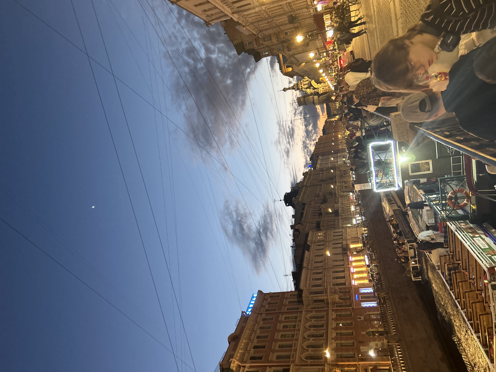 The image depicts a canal scene in a European city during twilight. The sky is a deep blue, with scattered clouds and a crescent moon visible. Buildings line the canal on both sides, showcasing ornate architecture with lit windows and streetlights. Several tour boats are present on the water, filled with people. One boat in the foreground is empty of passengers, with rows of wooden seats. On the right side of the canal, a pedestrian walkway is bustling with people, some of whom are looking towards the canal. The overall atmosphere suggests a popular tourist area enjoying an evening activity.