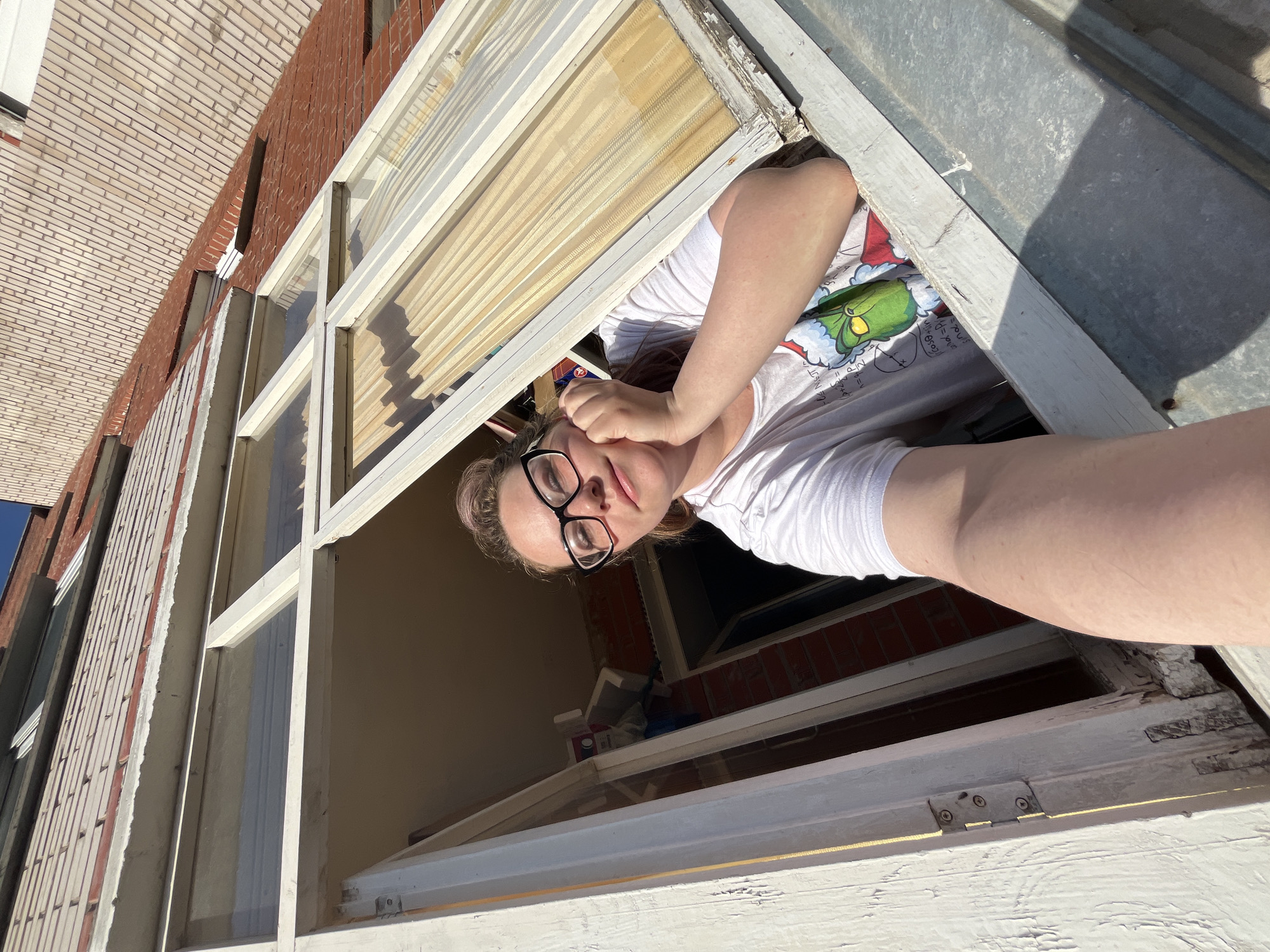 A woman with glasses and a white T-shirt with a Grinch image is looking down from an open window. The window frame is white and peeling. Outside, there is a brick building. Inside, there are painting supplies. The location is likely Chaikovsky, Russia.