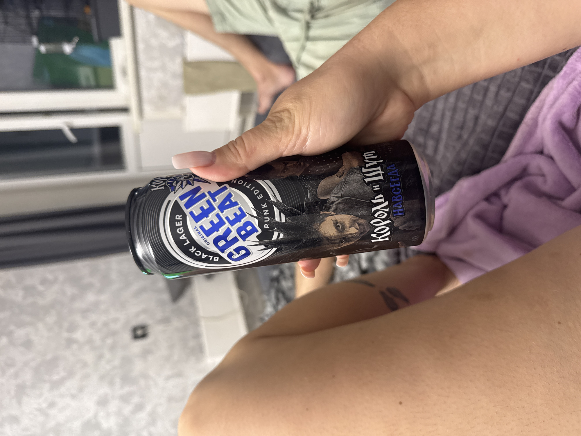 A person's arm and hand are in the foreground, holding a can of "Green Beat" black lager. The can has an illustration of a punk character and text in Cyrillic script. The background shows a bed with gray and purple bedding, and a window with blinds. The scene appears to be indoors, likely in a residential setting, with no discernible features placing it specifically in Tolyatti, Russia.