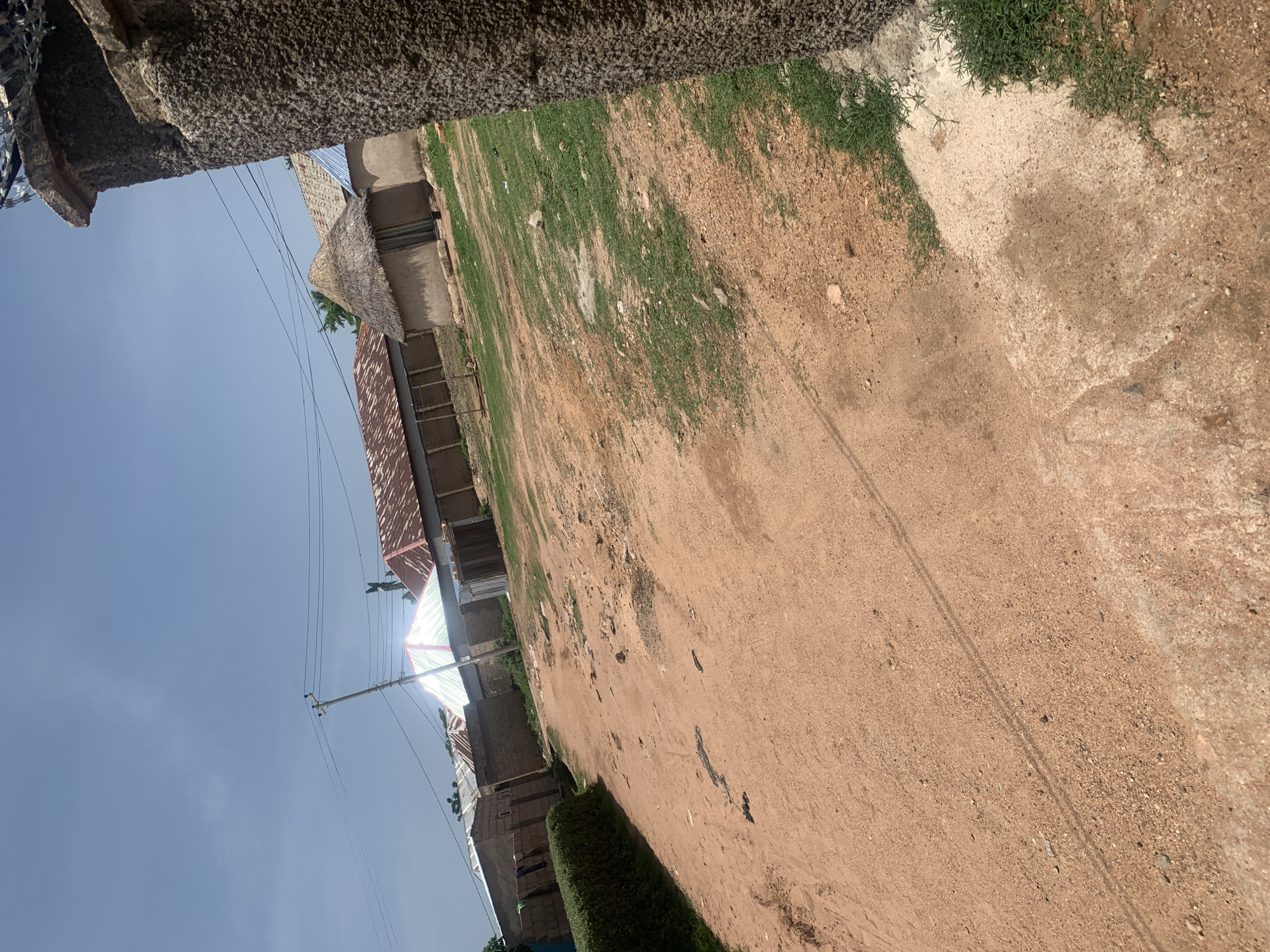 The image depicts a dirt road in the city of Jauro Yinu, Nigeria. The road is lined with various structures, including buildings with corrugated metal roofs, one with a thatched roof, and a conical structure with a reflective surface. There are also power lines with a utility pole visible. The environment appears to be a rural or semi-rural area, with dry earth and patches of grass. A rough stone pillar with barbed wire on top is in the foreground on the right, partially obscuring the view of the road and buildings. The sky is blue with some scattered clouds. There are no people visible in the image.