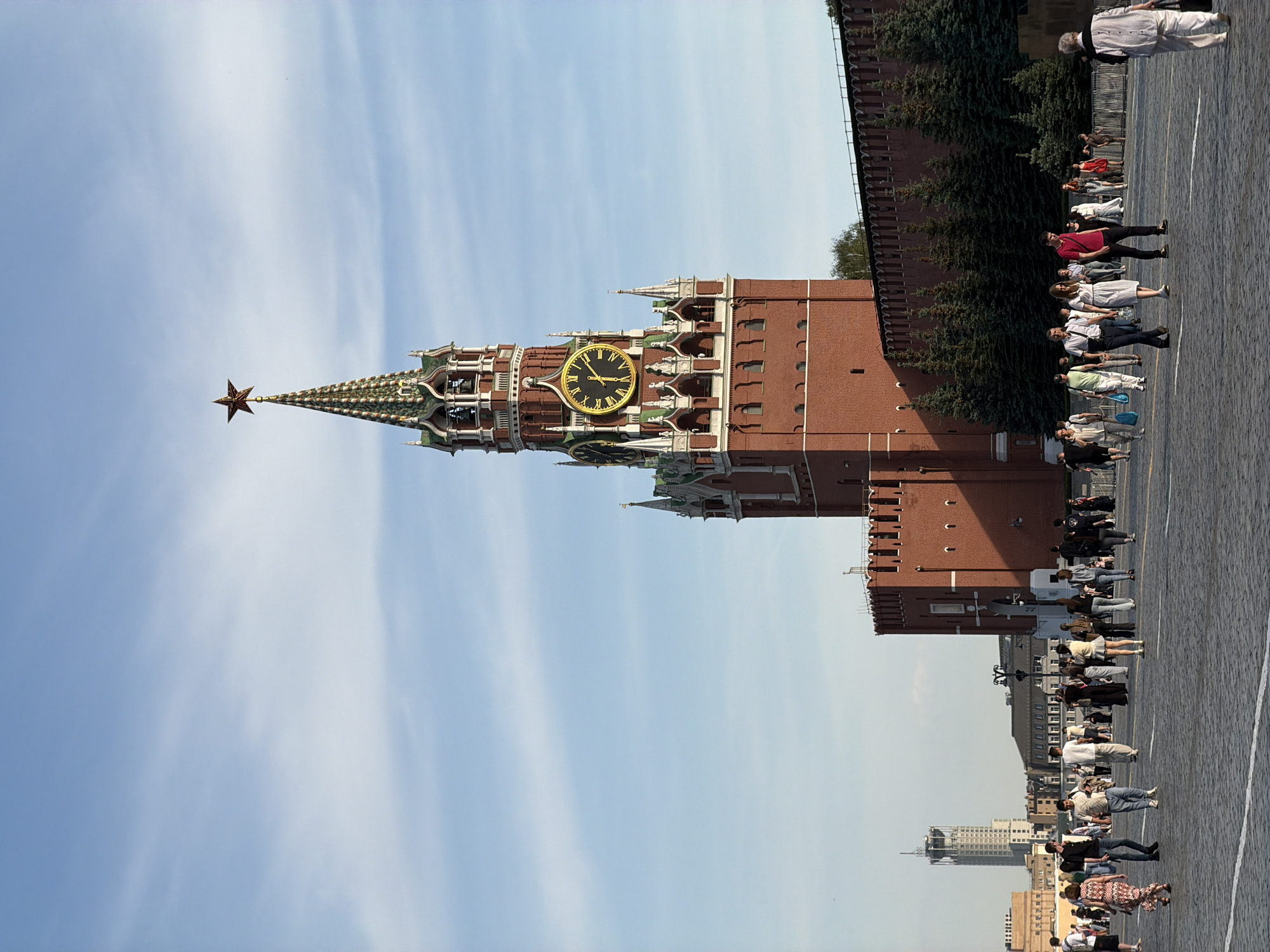 The image depicts the Spasskaya Tower of the Moscow Kremlin in Moscow, Russia. The tower, made of red brick, features a clock face and is topped with a red star. People are walking near the Kremlin walls. The sky is blue with white clouds. The tower is angled to the left of the frame. A modern building is visible in the background.