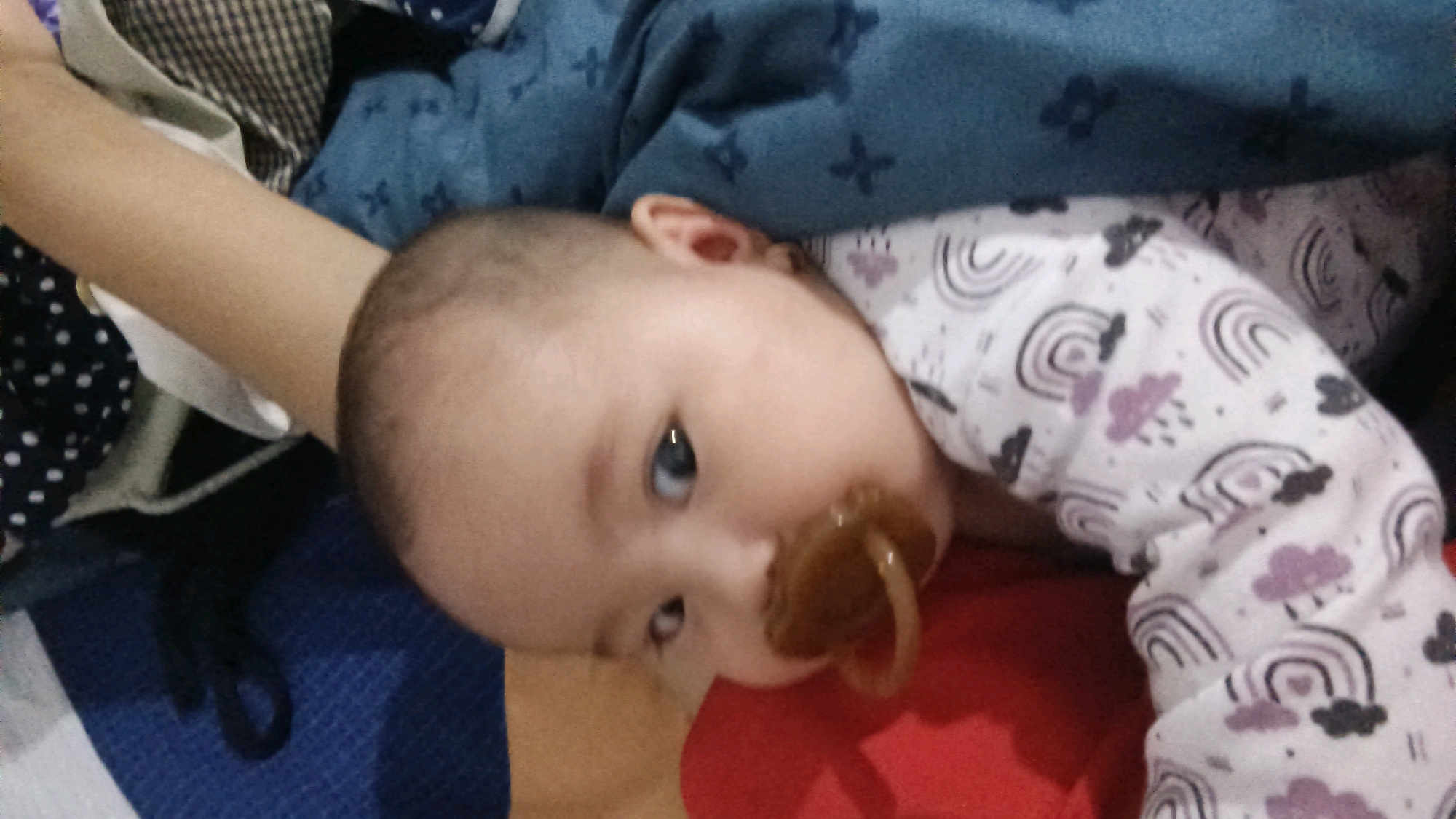 A close-up, top-down shot shows a baby in Donorejo, Indonesia. The baby is lying on its back, with its head tilted to the right. The baby is wearing a white pajama set with a rainbow and cloud pattern. The baby has a pacifier in its mouth and its eyes are open. The baby's left arm is visible, resting on a blue blanket with a cross pattern. The background is a mix of blue and brown fabrics. The lighting suggests an indoor setting, possibly a home.