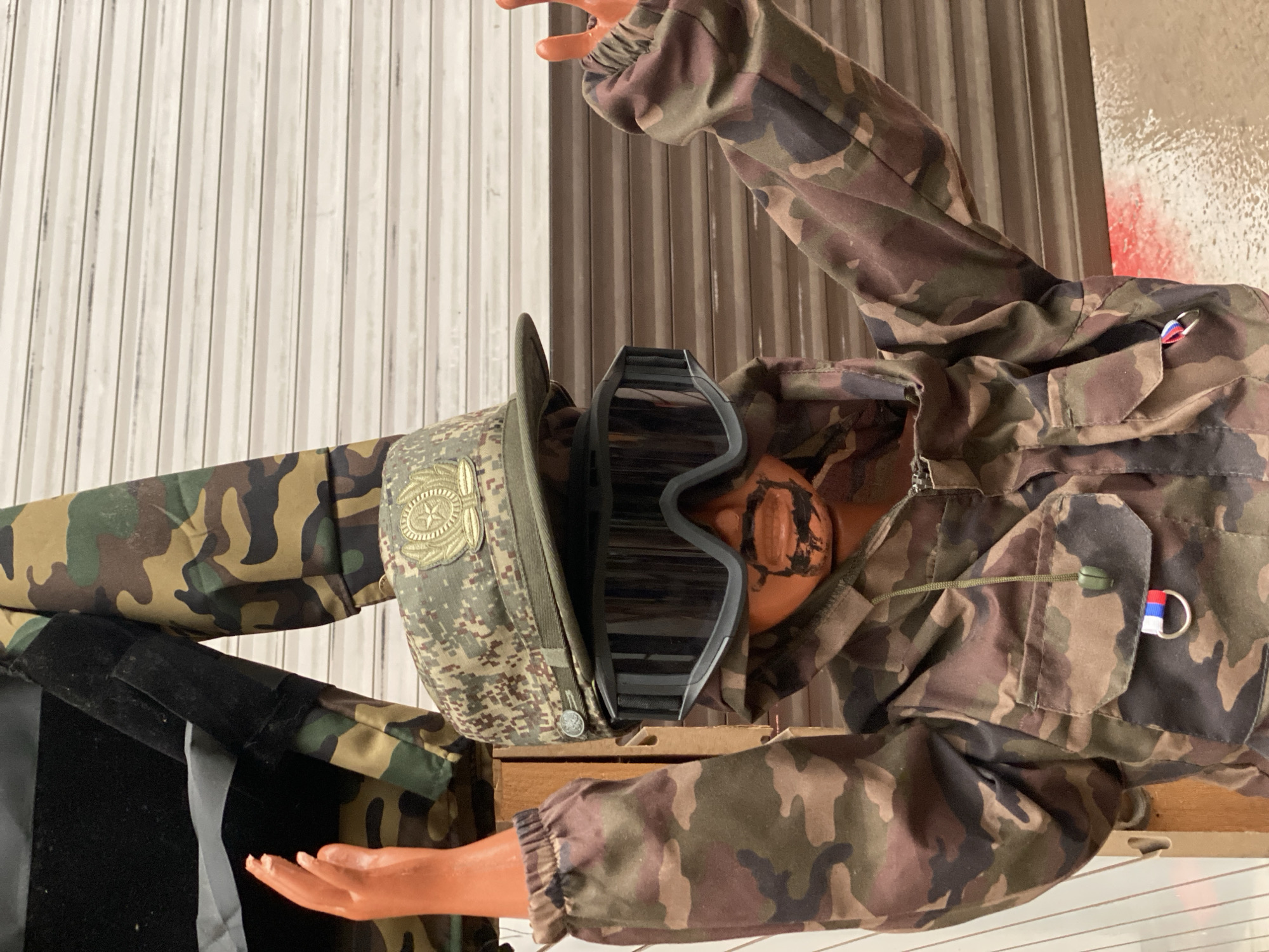 A mannequin dressed in a camouflage jacket and pants, a camouflage cap with a gold emblem, and large black goggles is standing with its arms raised. The mannequin has orange hands and a painted mustache and beard. A small Russian flag is attached to a ring on the jacket pocket. The background is a corrugated metal garage door. The scene appears to be outdoors, possibly in a yard or alley.