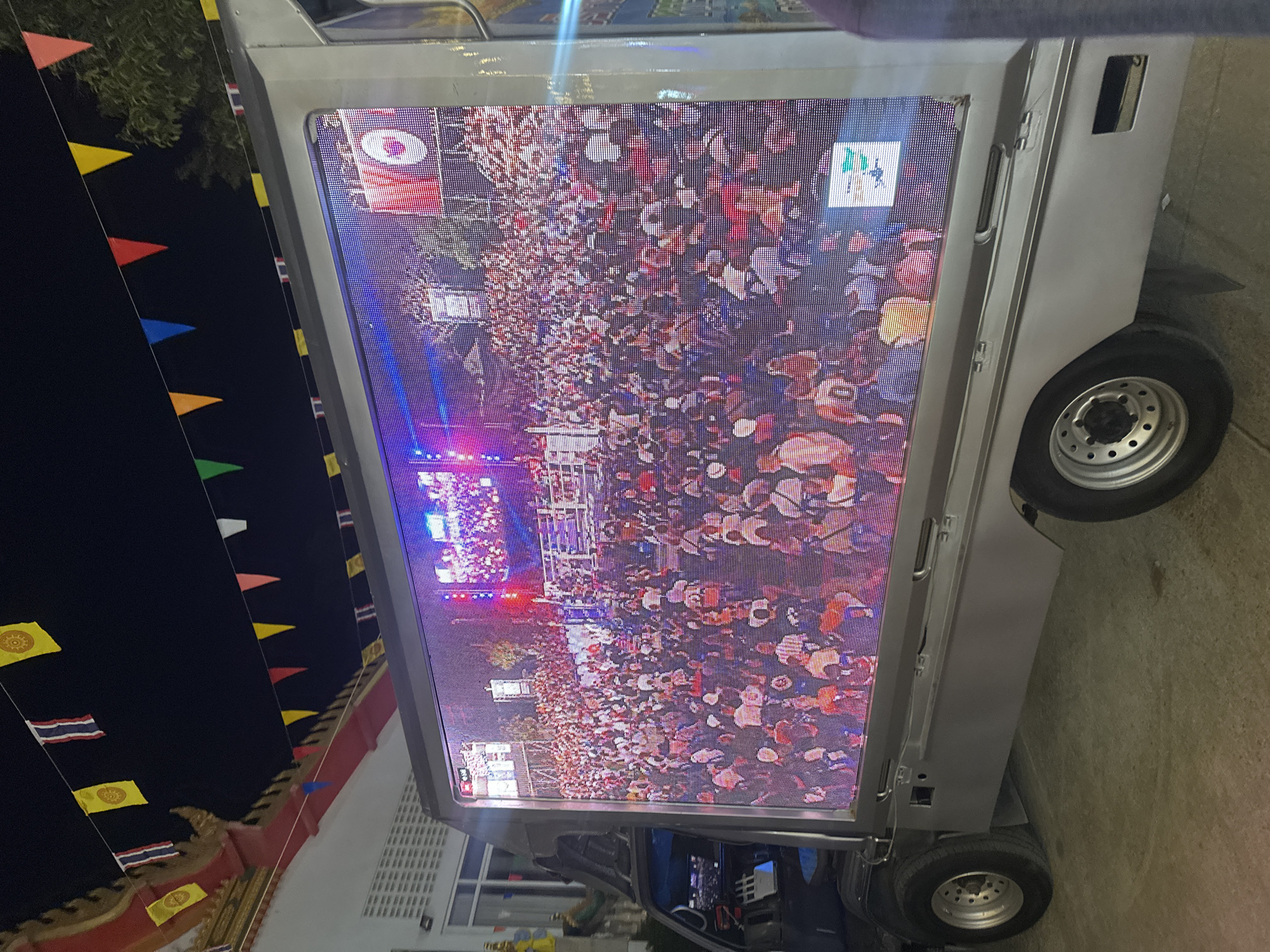 Here is a detailed description of the image:
The image shows a large digital display mounted on the back of a vehicle, possibly a small truck or van. The screen is displaying what appears to be live footage of a crowded event or gathering, possibly a concert or festival. The crowd is dense and mostly composed of individuals, with the stage lit up with lights.
The location is บ้านบางรักษ์, Thailand. This is inferred from the presence of Thai flags along with other triangular pennants near a building's roof line.