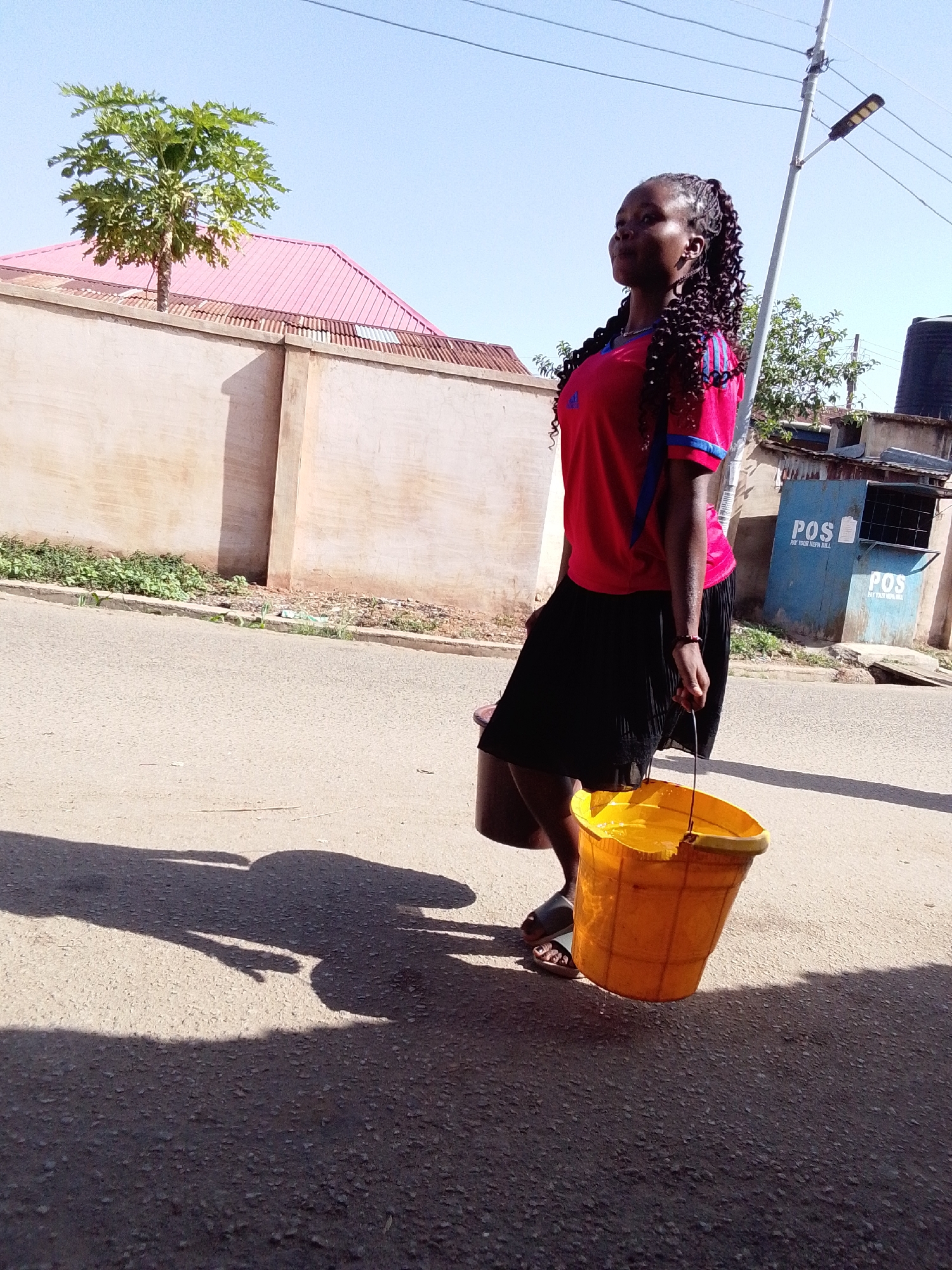 A young woman with braids is walking on a paved road, carrying a yellow bucket filled with liquid. She is wearing a red t-shirt with blue stripes on the sleeves and a black skirt, along with sandals. Behind her is a beige wall with a pink roof visible. A tree is growing behind the wall. To the right, a building with a blue facade labeled "POS" is visible. A light pole stands next to the building. The environment suggests a sunny day in a residential area. The city of Jos, Nigeria is not overtly indicated.