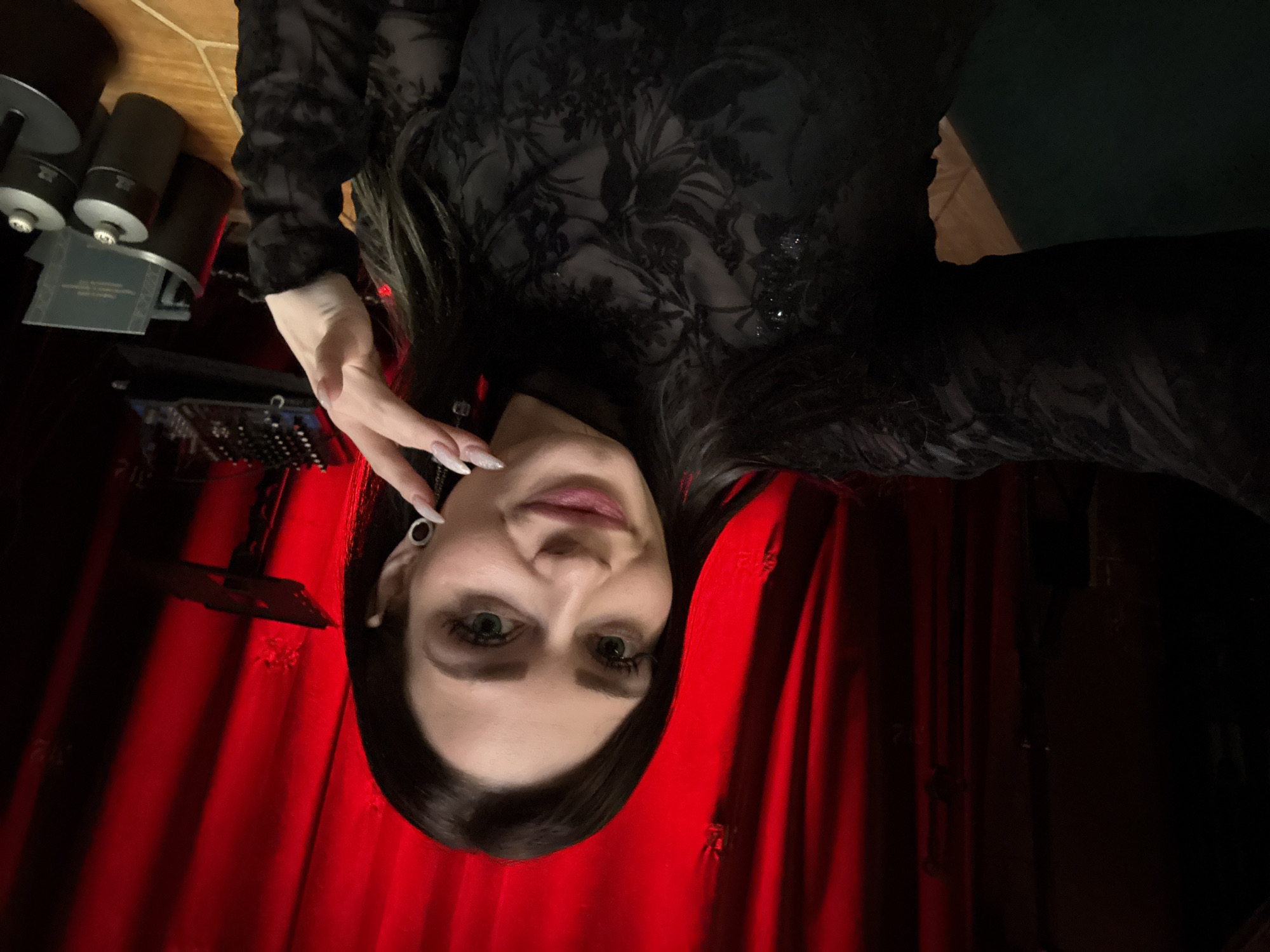 The image is a close-up shot of a woman with long, dark hair wearing a black, patterned top. Her complexion is fair and her eyes are green. She has elongated nails with a light-colored polish. Her left hand is raised near her face.
The background consists of red curtains or drapes. To the left of the frame, there is a collection of dark cylindrical objects, potentially lighting equipment, and an electronic device with various buttons and dials. There are no indications of the location being in Moscow, Russia.