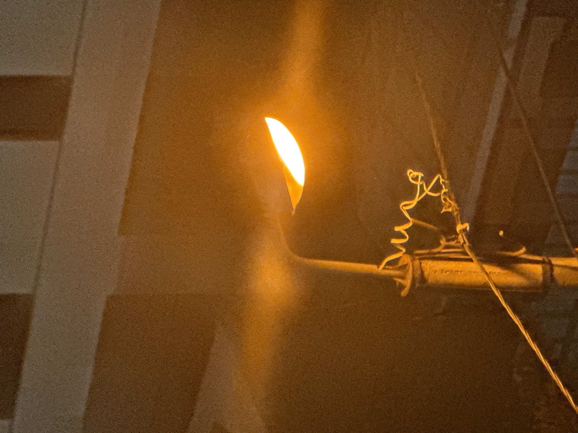 The image shows a street lamp with a bright orange glow, likely a sodium-vapor lamp, illuminating a textured, dark background that suggests a building facade. The lamp is mounted on a pole with a coiled wire attached. A thin wire runs diagonally across the lower part of the frame. The overall lighting and texture suggest the image was taken at dusk or night in an urban environment, potentially in Tbilisi, Georgia, given the context. No people or specific interactions are visible. The scene is static and depicts a common urban fixture.