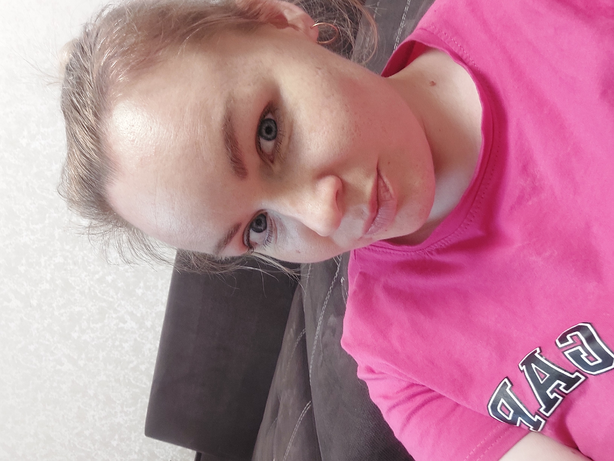 A young woman with fair skin and blue eyes is pictured in a close-up selfie. Her hair is light brown and pulled back. She is wearing a bright pink T-shirt with the letters "GAP" visible in reverse. She is sitting on a dark brown sofa. The background shows a white patterned wall. The location is Chaikovsky, Russia.