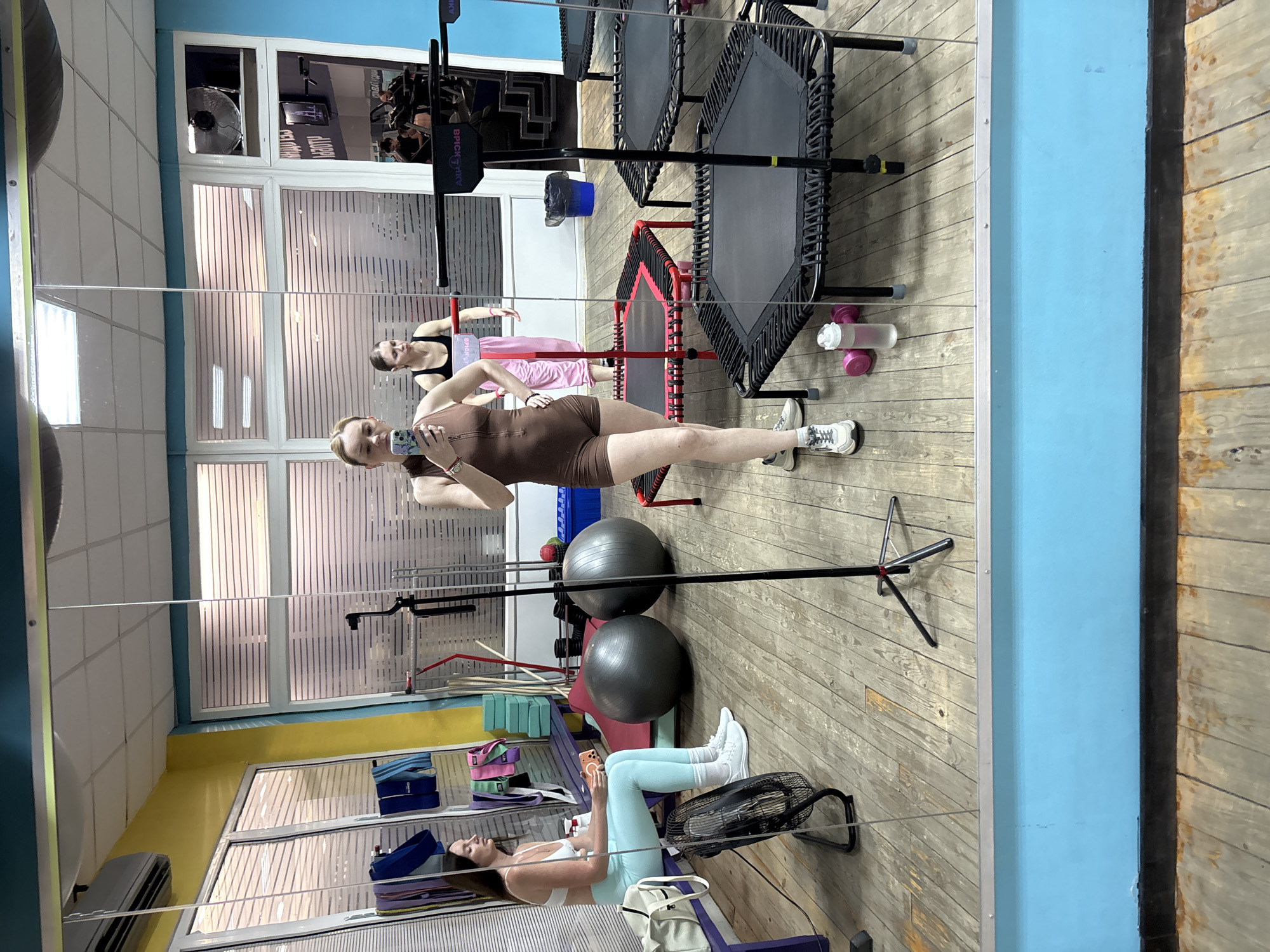The image captures a scene inside a fitness studio, likely located in Saint Petersburg, Russia, viewed through a reflection in a large mirror or glass partition. The flooring consists of light-colored wooden planks, complemented by walls painted in sections of bright blue and yellow, with some areas featuring white tiles.
Three women are present, all appearing to be engaged in or resting from exercise. In the prominent foreground reflection, a woman with blonde hair stands, dressed in a brown, form-fitting athletic unitard and white sneakers. She holds a smartphone with a colorful case, seemingly taking a photo or video, with one leg slightly bent. Behind her, partially visible, is another woman wearing a black top and light pink athletic pants, standing near a mini-trampoline. Further back and seated on the wooden floor, a third woman with long dark hair wears a white sports bra, light blue leggings, white socks, and white sneakers, absorbed in her smartphone. A black oscillating fan is positioned near her feet, and a light-colored bag rests beside her.
The studio is equipped with various fitness items, including several black and red mini-trampolines (rebounders), two large grey exercise balls, and colorful resistance bands stacked on a shelf. A pink and clear water bottle is visible on the floor.
Notable visible text includes a partial brand name "BRICKY" on a black vertical support bar, and unreadable Cyrillic text on a screen reflected in a distant window. The bright lighting and array of equipment suggest an active workout environment.