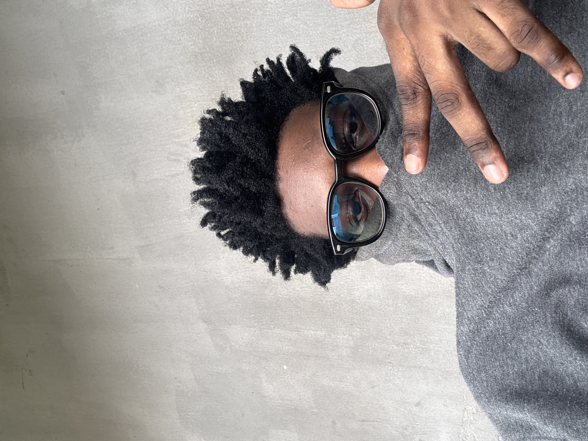 A person with dark skin and black curly hair is wearing glasses and a gray turtleneck. Their face is partially obscured by the turtleneck, with only their eyes and forehead visible. They are making a hand gesture, with their index and middle fingers extended and their thumb and ring finger bent. The background is a plain gray wall. The image appears to have been taken in Ihiagwa, Nigeria, though no specific landmarks are visible.