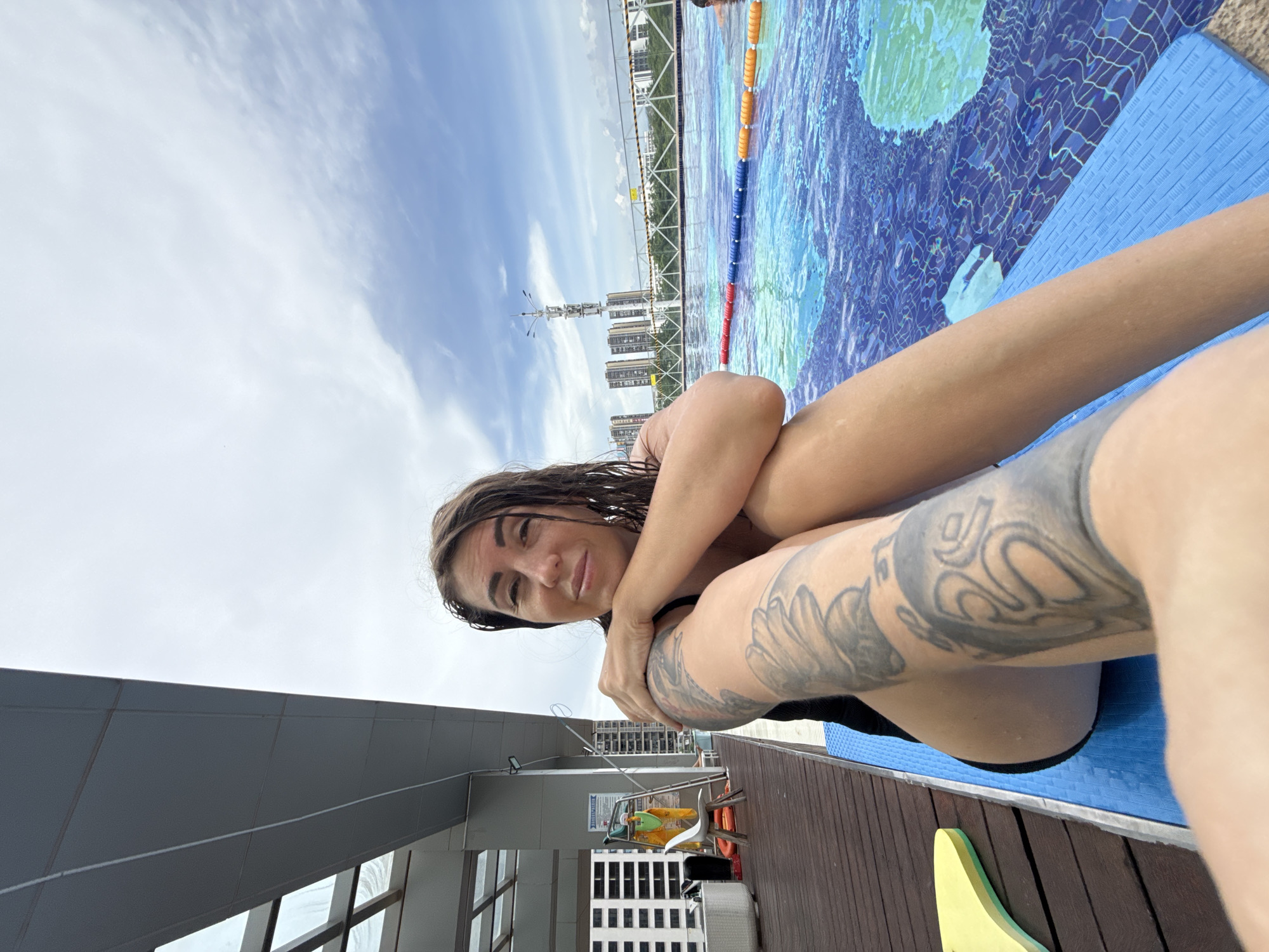 A woman with wet hair and tattoos on her left arm sits on a blue mat next to a swimming pool. The pool has lane markers, and there are buildings in the background. The scene appears to be an outdoor swimming pool in a city, possibly Huizhou, China, given the architectural style of the buildings and the presence of high-rise structures. The sky is cloudy, suggesting it is daytime. There are several objects on the wooden deck to the left of the woman, including a white chair, a yellow life preserver, and some other pool equipment.