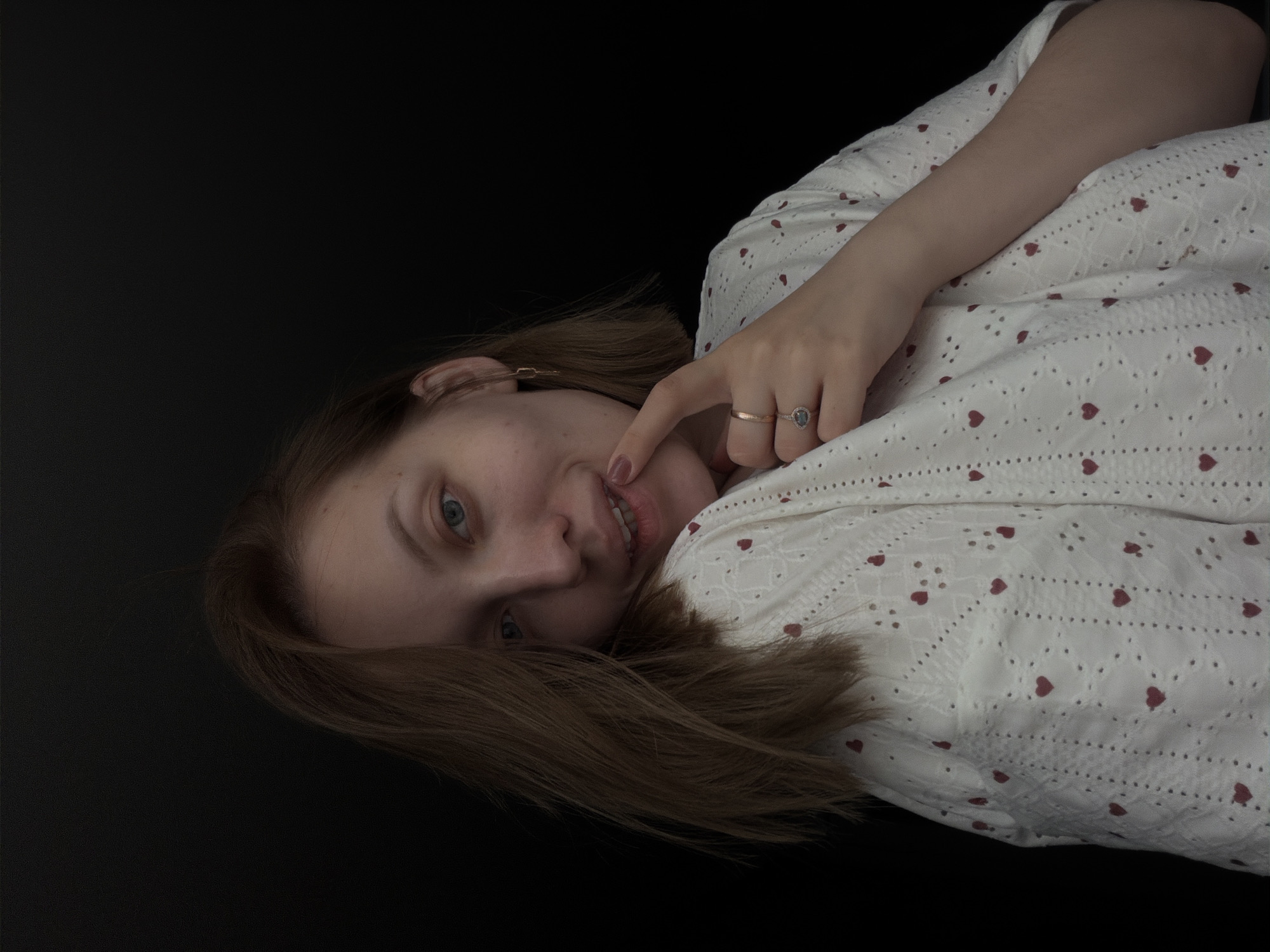 A young woman is depicted lying on her side, with her head tilted and looking directly at the camera with a soft, playful smile. The image itself is rotated 90 degrees clockwise, making her appear to be lying horizontally against a dark background. She has light brown to auburn hair that flows around her head and fair skin. Her eyes are blue-grey, and her lips are slightly parted, revealing a hint of teeth, while her right index finger gently touches her lower lip.
She is dressed in a white top adorned with small red heart patterns and delicate eyelet embroidery. On her right ring finger, she wears two rings: one a plain gold band and the other a gold band set with a dark, oval gemstone. A small, golden hair clip is visible tucked into her hair near her right ear.
The background is a solid, dark black, suggesting an indoor setting with a plain backdrop, which emphasizes the subject. The lighting is soft and even, highlighting her features and the texture of her clothing against the dark void. There are no specific location cues visible within the image itself, beyond the contextual information that it was taken in Reshetnikovo, Russia. No visible text is present.