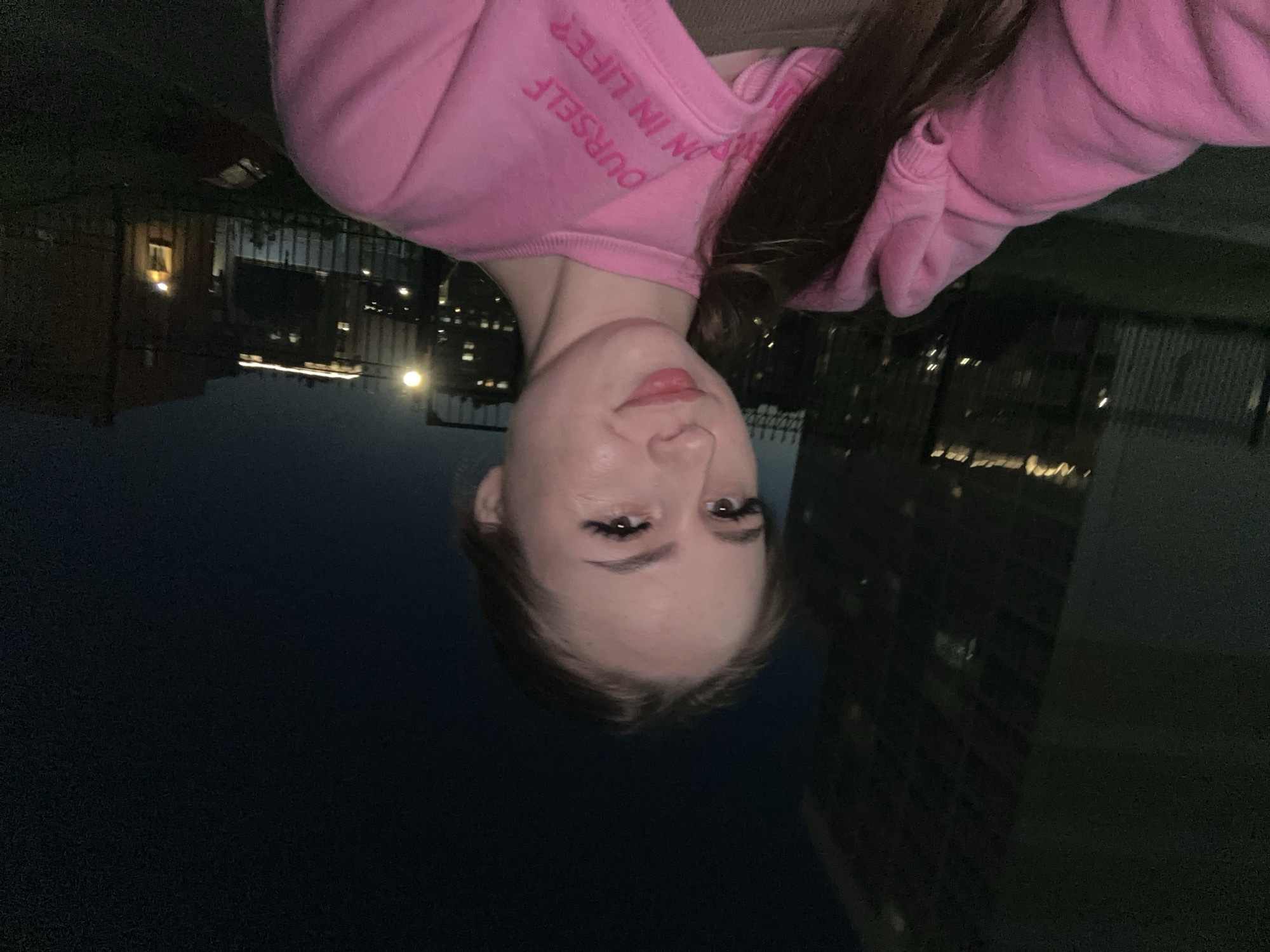 A close-up selfie captures a person, likely female, facing the camera with a neutral to slight smile. Her fair skin, defined eyebrows, and long eyelashes are prominent, along with light pink or nude lipstick on her lips. She wears a pink hoodie with text printed on it; visible fragments include "YOURSELF" and "ON IN LIFE". Long, dark hair frames her face and extends over her shoulders. The background is dark, indicating it is nighttime. Distant city lights from buildings and possibly streetlights are visible, suggesting an urban setting. Faint outlines of structures and what appears to be a fence or railing are also discernible in the low light.