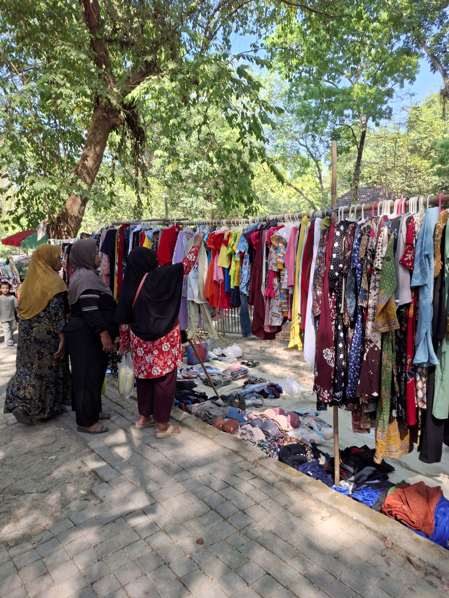 In Tawun, Indonesia, an outdoor market is in full swing. A rack of colorful clothing hangs on display, featuring dresses, shirts, and other apparel. Several women browse the merchandise, their faces partially obscured by headscarves. A young child stands nearby, observing the scene. Piles of clothing are spread on the ground, indicating a bustling marketplace. Trees provide shade, casting dappled shadows on the paved walkway. The atmosphere is lively, with the presence of multiple vendors and shoppers.