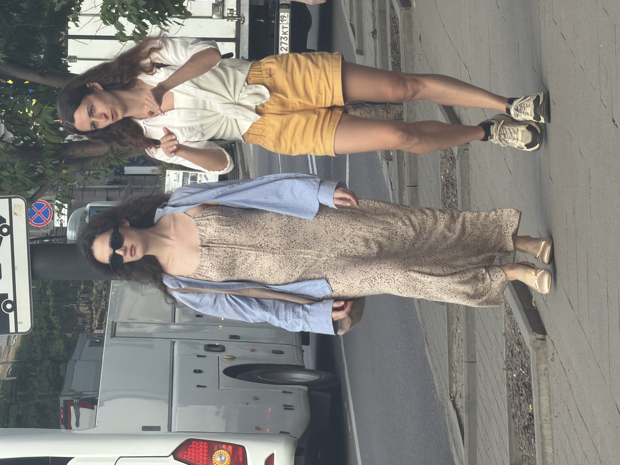 Two women walk on a paved sidewalk in the city of Krasnoyarsk, Russia. The woman on the left, with long, dark, curly hair, wears a beige, patterned, sleeveless maxi dress, an open blue and white striped shirt, and beige flats. She has on black, oval sunglasses. A tan crossbody bag is slung over her left shoulder. The woman on the right, with long, dark, wavy hair, wears a white, tied-front short-sleeved shirt, yellow shorts, and black and white sneakers with black socks. She is gesturing with her hands. Behind them, a light grey bus is parked on the left side of the street. A white truck with a license plate reading "273KT 19" is visible on the right. Trees line the background, and street signs are visible above the bus.