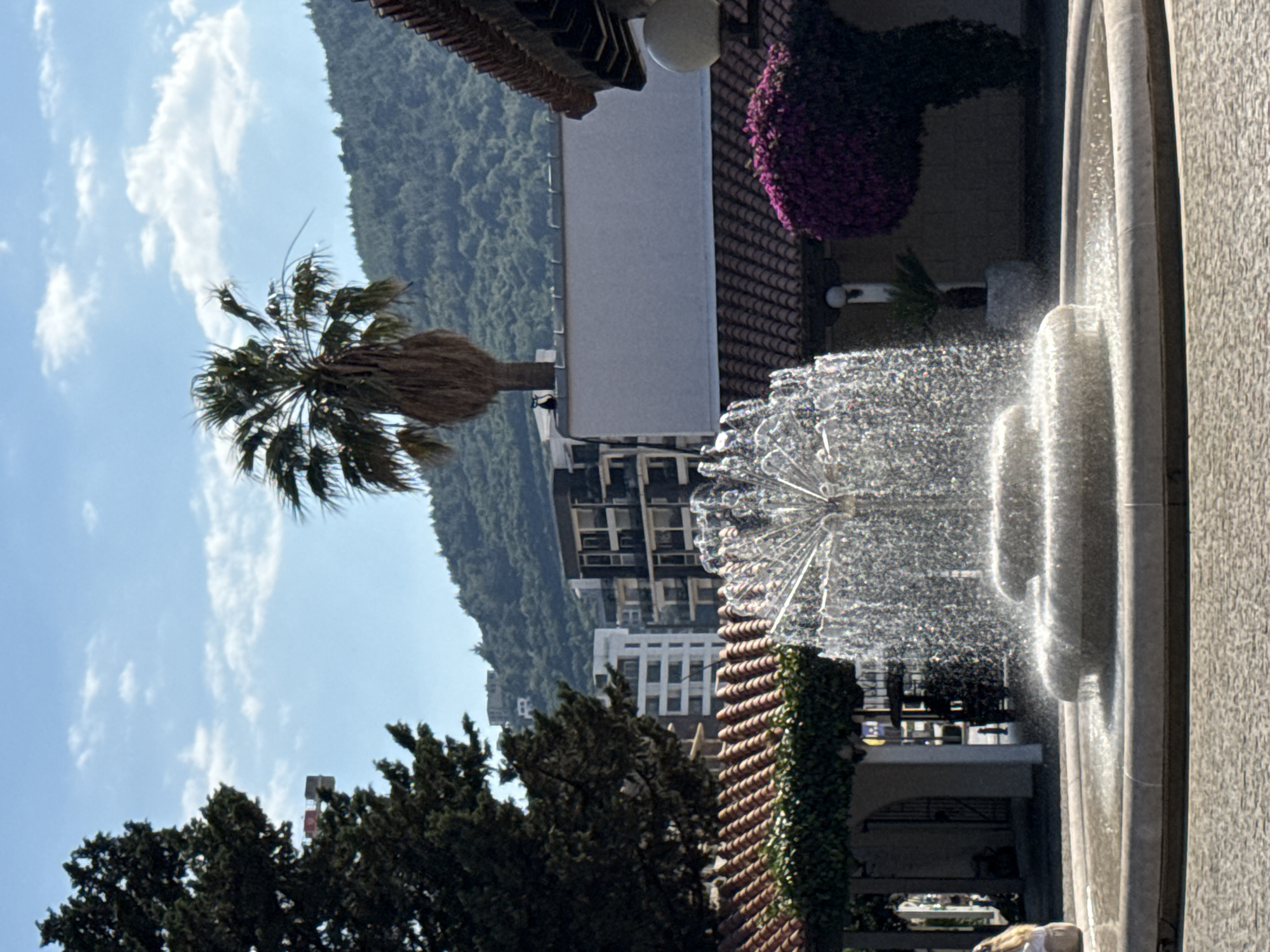 The image shows a circular stone fountain in a paved plaza, with water spraying upwards and outwards in a fine mist. Behind the fountain, there is a building with red tiled roofs and some greenery. Further back, there are several modern buildings, including a tall white one and darker ones with balconies, situated on a tree-covered hillside. A palm tree stands between the fountain and the buildings, and a large evergreen tree is visible on the left. The sky is blue with scattered white clouds. The scene appears to be in Budva, Montenegro, possibly near the Old Town or a waterfront area, given the architectural style and the presence of palm trees and the hilly backdrop.