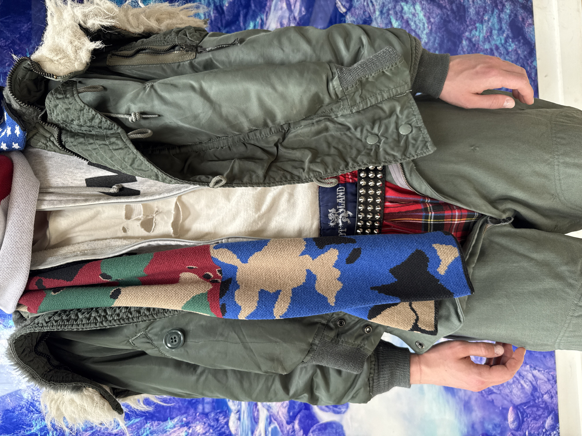 The image depicts a person wearing several layers of clothing. The outer layer appears to be an olive green parka with a fur-lined hood. Underneath, there are visible layers of clothing including a grey sweatshirt, a white t-shirt, a plaid garment, and a colorful camouflage patterned scarf. A studded belt is also visible. The person is wearing matching olive green pants, and their hands are visible on either side of the image. The background appears to be a mottled blue and white surface.
