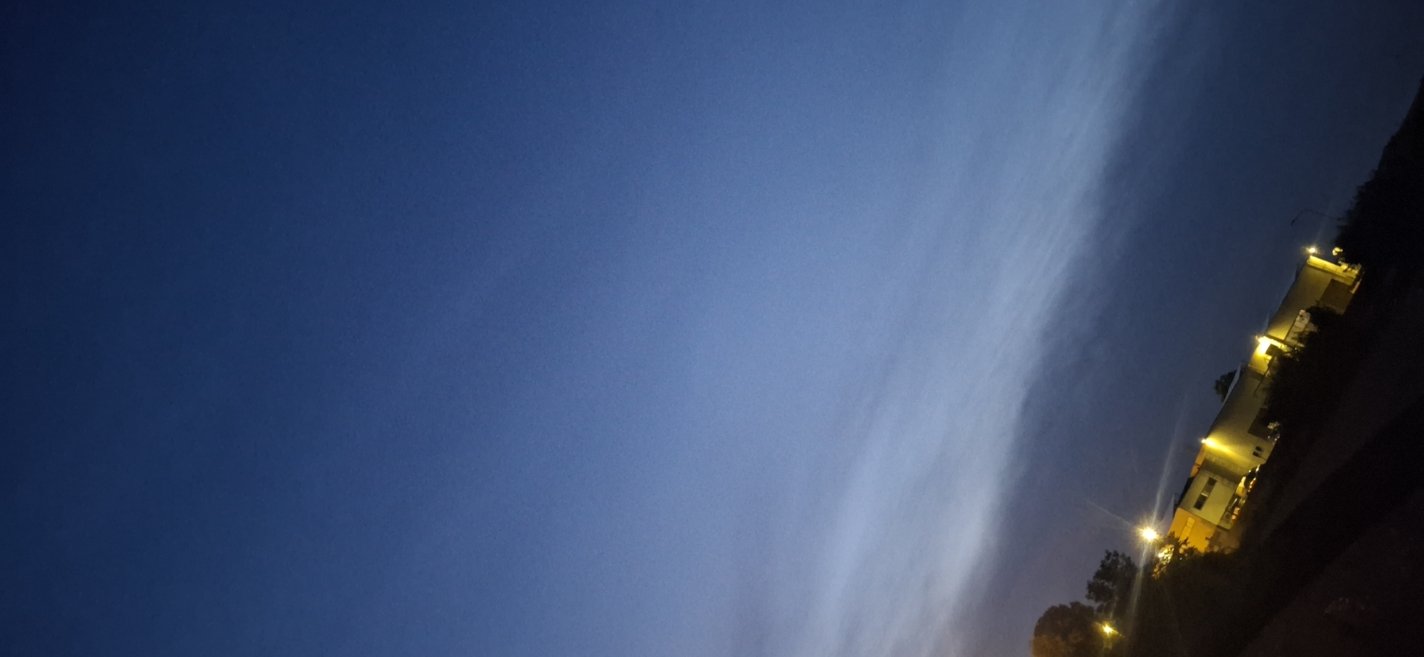 A diagonal shot of an industrial-style building at dusk or night in Sant'Egidio alla Vibrata, Italy. The sky is a deep, clear blue that fades to a lighter shade near the horizon, with faint wispy clouds visible. The building, illuminated by bright yellow lights, appears to have a light-colored facade and a dark roofline. Several windows are visible, some with lights on. Streetlights are also present, casting pools of light. Trees and vegetation frame the building. The overall impression is one of a quiet, commercial area as day turns to night.