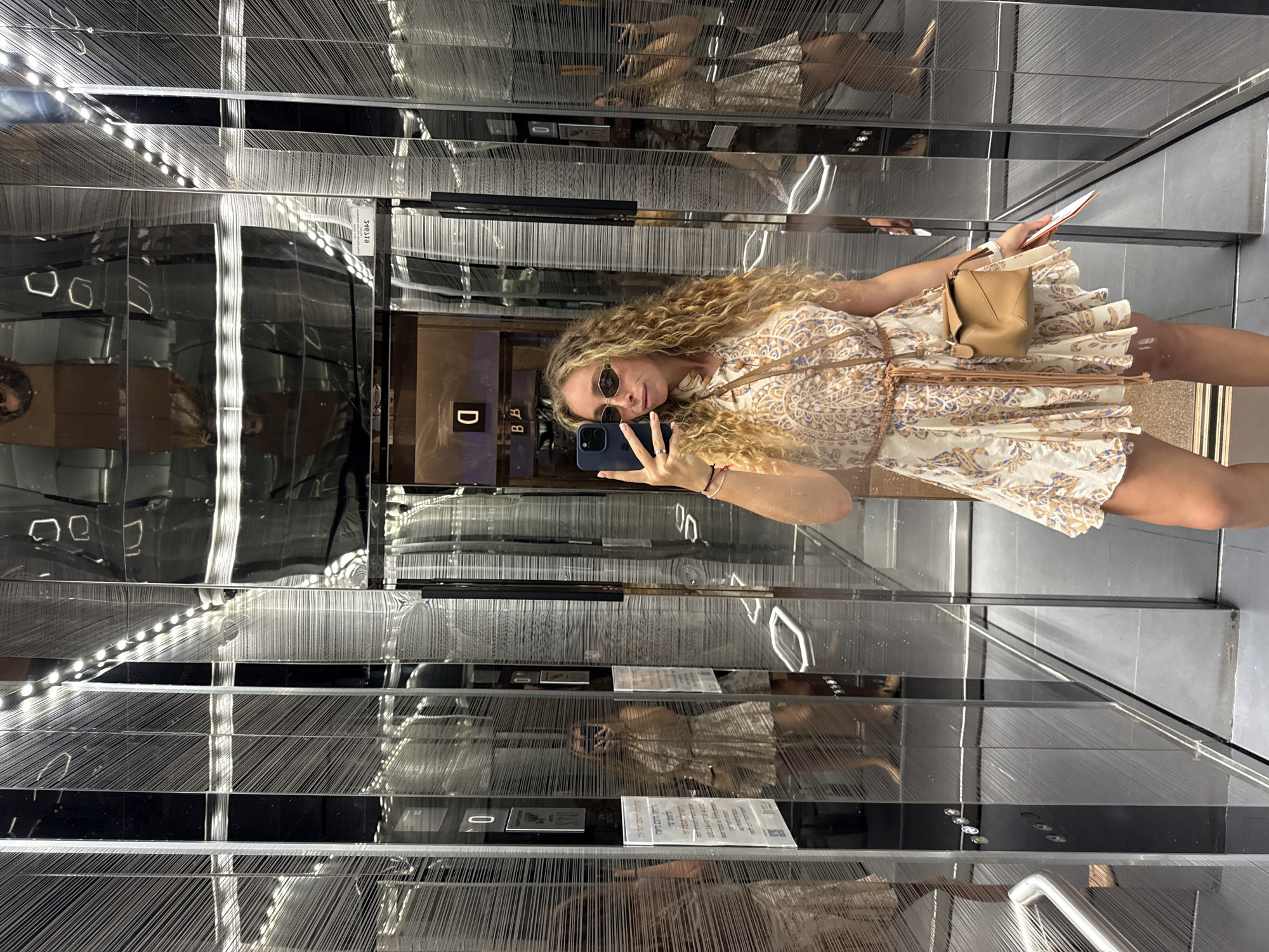 A young woman with curly blonde hair and sunglasses is taking a selfie in an elevator in Tel-Aviv, Israel. She is wearing a patterned dress and a small brown crossbody bag. The elevator has mirrored walls with a textured pattern. Reflections of the woman and other parts of the elevator are visible in the mirrors. The elevator appears to be in a modern building, possibly a hotel or residential complex.