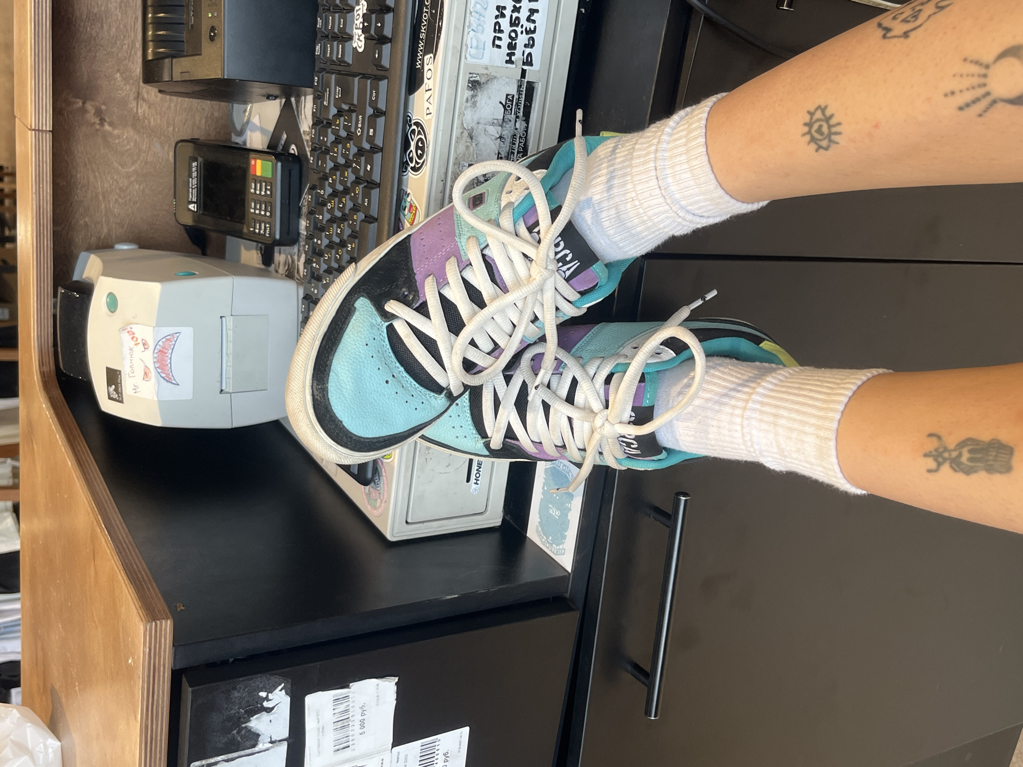 The image shows a pair of legs wearing white socks and pastel-colored sneakers, presumably in the city of Krasnoyarsk, Russia, given the presence of Cyrillic text and a "5,000 руб" label, which is likely related to the Russian ruble currency. The legs are positioned on a dark-colored desk or counter. The desk has a light brown wooden edge and is cluttered with various items, including a keyboard, a credit card terminal, a label printer with a cartoon sticker on it, and other unidentifiable objects. The legs have tattoos on the visible parts of the lower arms.