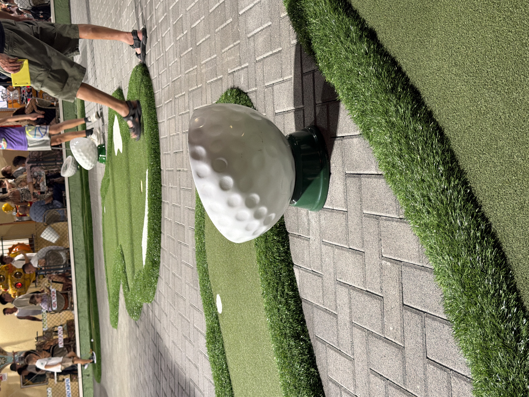 The image shows a mini-golf course in Gatteo, Italy, with a large decorative golf ball sculpture in the foreground. The golf ball is white with dimples and sits on a green base. The ground is paved with grey interlocking bricks arranged in a herringbone pattern. Green artificial turf borders the brick path and forms the mini-golf greens, which have white holes. In the background, several people are visible, some playing mini-golf and others standing around. One person on the right, wearing khaki shorts and sandals, is walking away from the camera. The overall setting appears to be an outdoor entertainment area, possibly a resort or amusement park, given the casual attire of the people and the recreational nature of the activity. The lighting suggests it is daytime, with some shadows indicating sunlight.