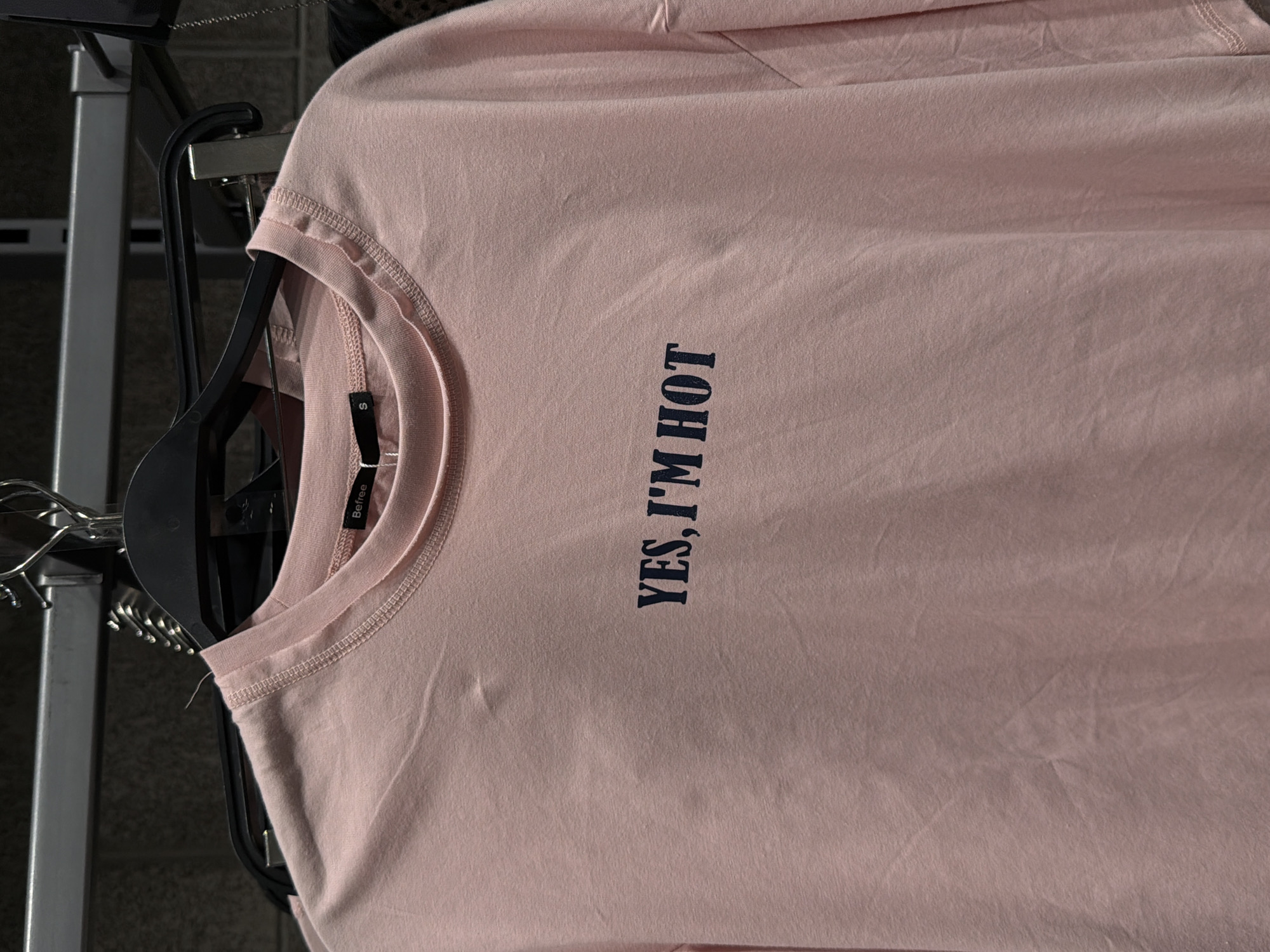 A light pink t-shirt with the words "YES, I'M HOT" printed in navy blue text is hanging on a hanger. The t-shirt has a tag with the brand name "Befree" visible. The background shows a clothing rack and a wall. The location is identified as a Befree clothing store in Moscow, Russia.