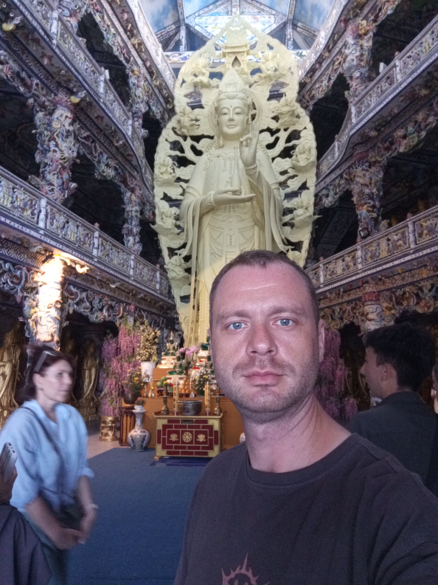 A man is taking a selfie in a dimly lit, ornate temple. In the background, a large, pale statue of a deity stands behind him. The man is wearing a dark t-shirt and has short, brown hair and blue eyes. To his left, a woman in a light blue shirt is visible, looking away from the camera. The temple's interior is richly decorated with colorful, intricate carvings and architectural elements. Further in the background, an altar with various offerings can be seen. The overall atmosphere is solemn and reverent. The location is identified as a temple in the city of Xuân Thành, Vietnam.