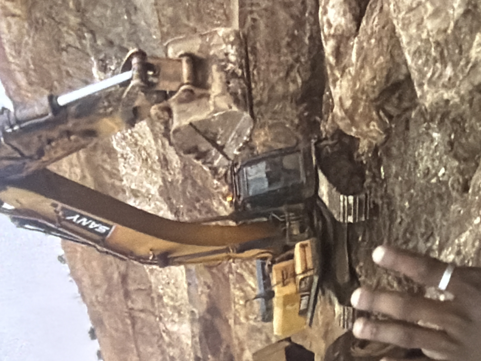 An excavator with the brand name "SANY" on its arm is visible in a quarry. The excavator is yellow and black, with a large bucket filled with soil. In the foreground, a hand with a ring on its finger is partially visible, with three fingers extended. The background consists of a barren, rocky landscape. The scene appears to be an active construction or mining site.