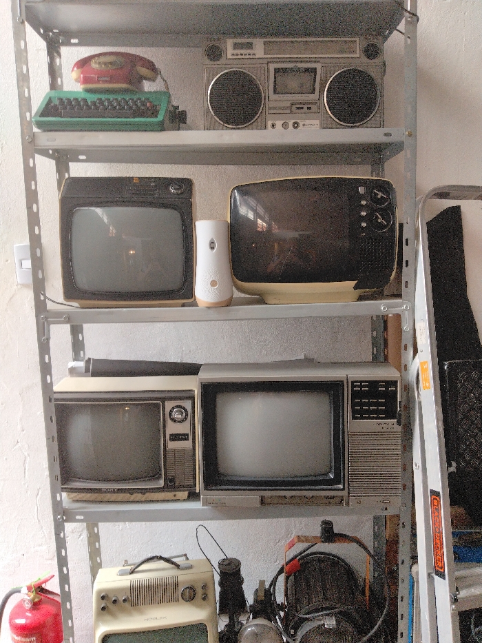 The image displays a multi-tiered metal shelving unit filled with a collection of vintage electronic devices, possibly in a storage room or garage.
On the **top shelf**, from left to right, there is a red rotary telephone, a green manual typewriter, and a silver-colored boombox (radio cassette player) with prominent speakers and central controls.
The **second shelf** holds two portable television sets and a modern air freshener dispenser. On the left, there's a dark-colored portable TV with a light beige base and a reflective screen. To its right, a white cylindrical air freshener dispenser stands between the two TVs. Further right is another dark-colored portable TV, also with a light beige base and a reflective screen, featuring control knobs on its right side.
The **third shelf** contains two more portable television sets. The one on the left is a lighter beige or cream color with a silver trim and a black screen. The word "SONY" is visible on its front panel. To its right, another light grey or silver-colored portable TV has a wide screen and a panel of buttons and a grille on its right side. A dark roll of material or a poster is lying horizontally behind these two TVs.
The **bottom section** of the image, below the third shelf, reveals a cluttered storage area. On the far left, a red fire extinguisher with some partially visible text is present. In the center, a cream-colored portable radio or small TV with a handle on top is visible, with the brand name "NOBLEX" clearly readable on its front. To its right, various wires, a black light fixture or stage light, and other mechanical parts are stored.
To the **far right** of the entire shelving unit, a metallic step ladder or similar ladder is leaning against the wall, with the brand name "BLACK+DECKER" visible on a sticker on its side. The background wall is painted a light color, possibly white, and has a textured finish. The overall impression is a collection of retro electronics meticulously stored.