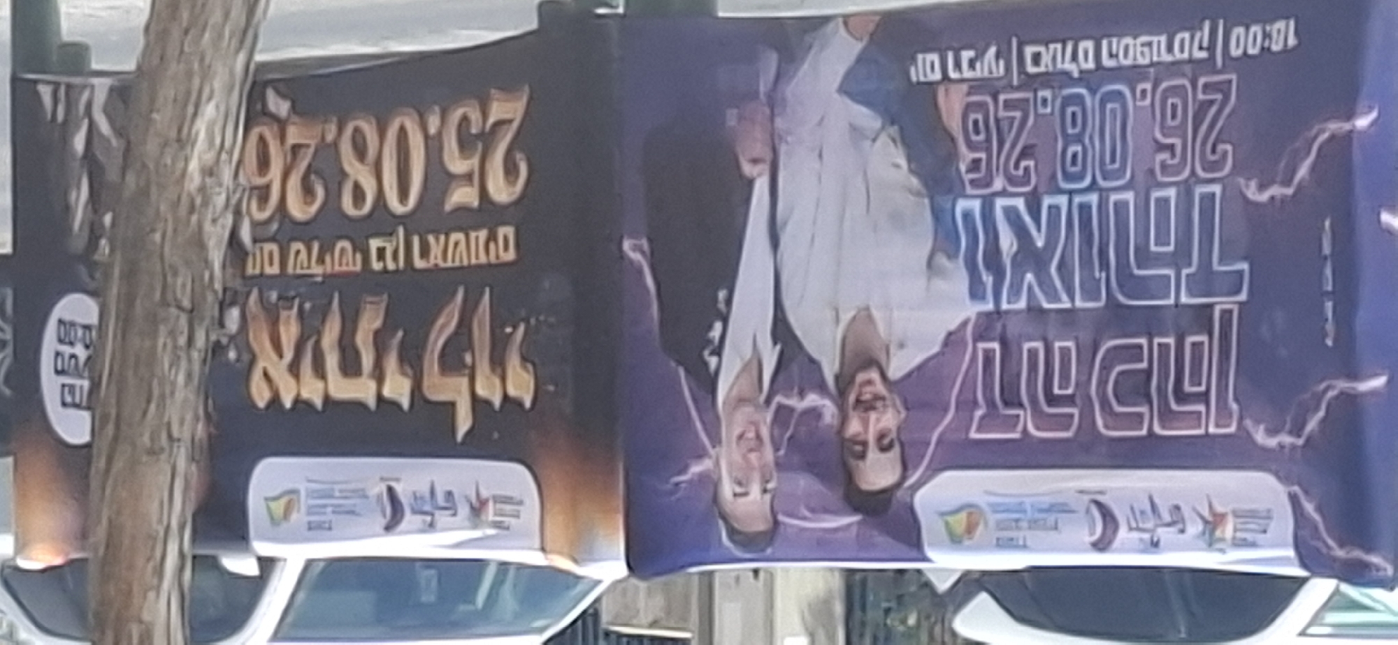 The image shows two banners displayed on the side of vehicles parked on a street, likely in Yerocham, Israel. The banner on the left features two men, one in a white shirt and the other in a suit. Large Hebrew text above them reads "Dedi Kah" and "and Yohid". Below their images, the date "26.08.26" is prominently displayed, along with smaller text indicating "Wednesday | Sports Hall | 18:00". The banner on the right has a dark background with large Hebrew text that appears to say "I Will Be My Own" or similar phrasing. Below this, it reads "Wednesday Ben R'ashem" and the date "25.08.26". A circular emblem is visible on the far right of this banner. The banners are set against a backdrop of trees and buildings, suggesting an outdoor urban environment. The lighting suggests it is daytime.