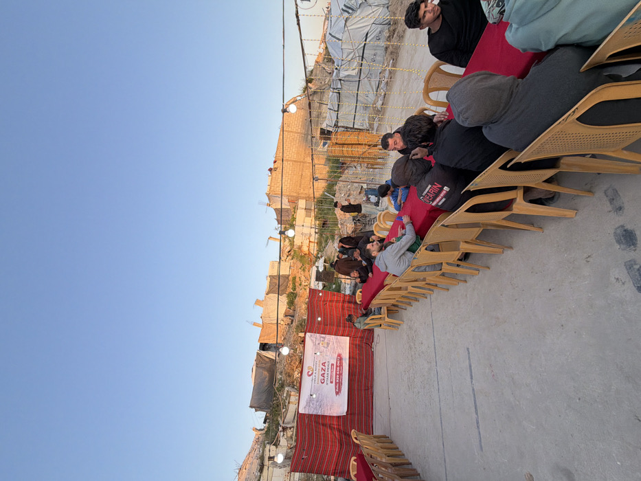 The image depicts an outdoor gathering set on a concrete ground surface in Jabalia, Palestinian Territory, under a clear, light blue sky. In the foreground, multiple long, narrow tables draped with red tablecloths are arranged parallel to each other. Numerous light brown plastic chairs are positioned around these tables. Approximately two dozen men are visible, predominantly seated at the tables, with some standing in the background.
To the left, a white banner with a red border and heart logo is prominently displayed against a red and white striped fabric. The banner reads "HELP A NEEDY," "GAZA," "#IFTAR MEALS," and includes a phone number: "Info: 939-636-0672." String lights with exposed white bulbs are suspended overhead, illuminating the area. The background reveals a landscape of severe structural damage and makeshift constructions. Rubble, damaged concrete structures, exposed rebar, and temporary shelters made from tarpaulins and various materials are visible on an elevated, uneven terrain. Additional temporary structures or stacked materials are present on the right side of the frame. The men at the tables appear to be waiting or conversing, while a few individuals in the mid-ground are standing near the damaged structures.