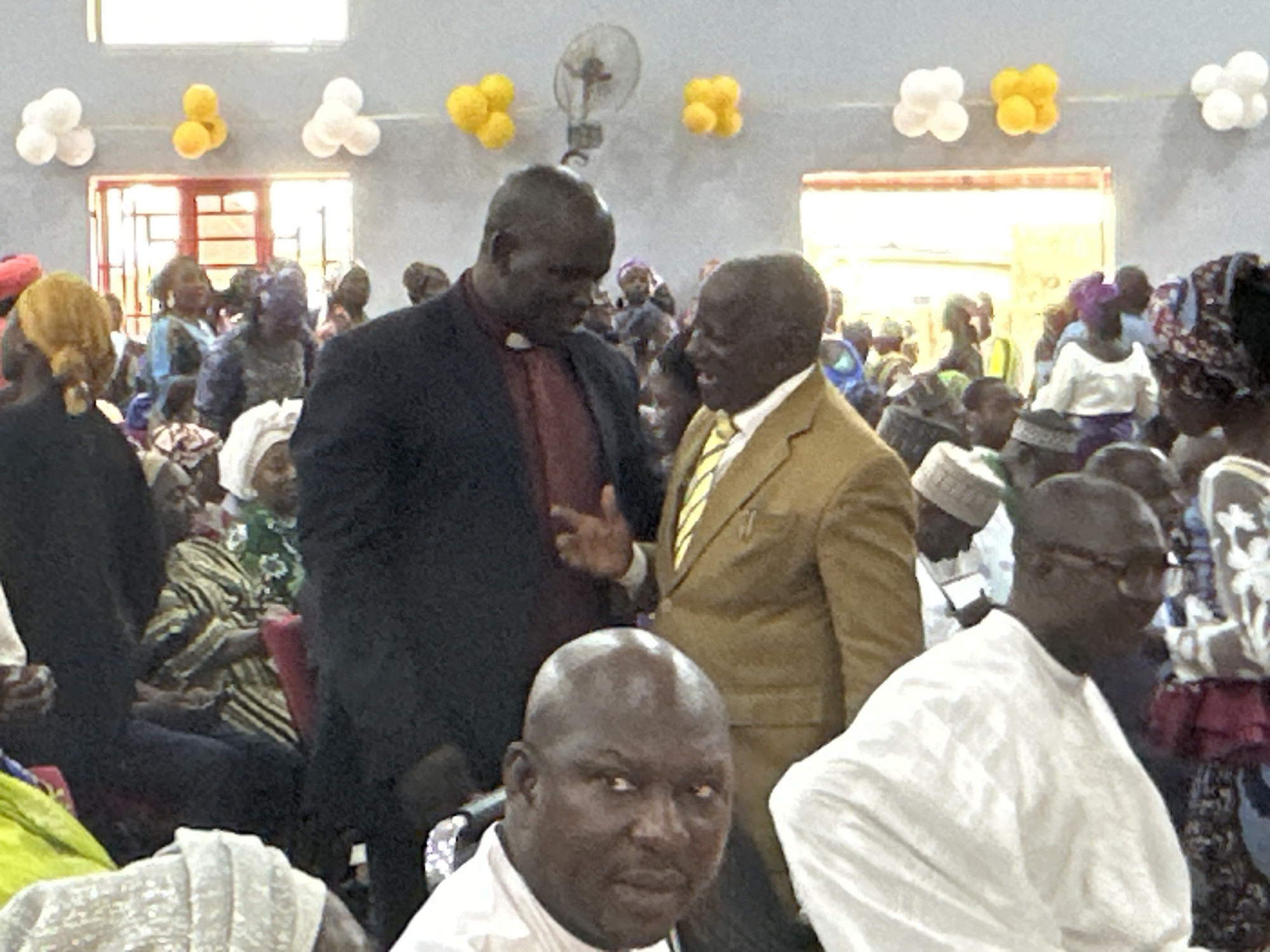 The image shows a gathering of people indoors, potentially in Goni Gara, Nigeria. Two men in the center are engaged in conversation. One man wears a dark suit with a clerical collar, indicating he may be a religious figure. The other man wears a light brown suit with a yellow tie. The crowd includes men and women of various ages, with many wearing traditional African clothing and head coverings. In the background, decorations resembling balloons are affixed to the wall. A ceiling fan is also visible. The lighting is somewhat muted, suggesting indoor conditions.