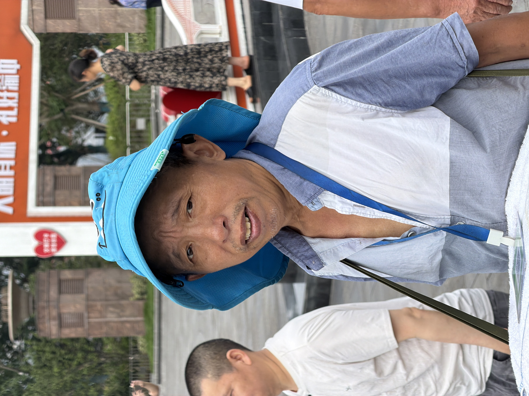The image shows a medium shot of an elderly Asian man wearing a blue bucket hat, a light blue and white collared shirt, and a blue lanyard. He has short, graying hair and appears to be looking at the camera with a slight smile. His left shoulder and chest are visible, along with the strap of a bag that is slung across his body. To his left, a portion of another man's head and shoulder is visible, wearing a white t-shirt. In the background, other people are visible, including a woman in a patterned dress. There is a building with an orange sign that reads "大明街·礼好满中" which is consistent with being in Yuzhong District, China. The scene appears to be outdoors, possibly in a public square or street.