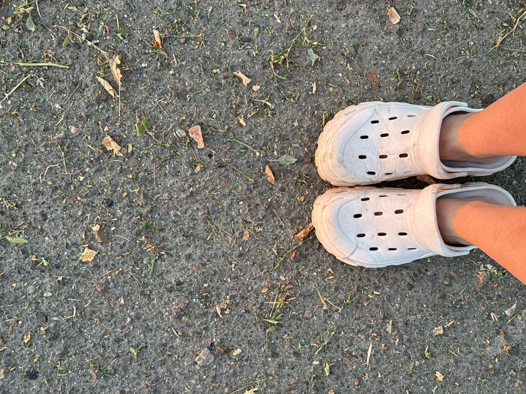 The image shows a top-down view of a person's feet wearing white Crocs on an asphalt surface. The asphalt is scattered with small pebbles, dirt, and dried leaves. The lighting suggests it is either early morning or late afternoon, casting an orange glow on the person's legs. The scene is consistent with a street or sidewalk in Aksay, Kazakhstan, where such surfaces and debris are common. There are no other individuals or significant objects present in the frame. The focus is solely on the footwear and the immediate ground.