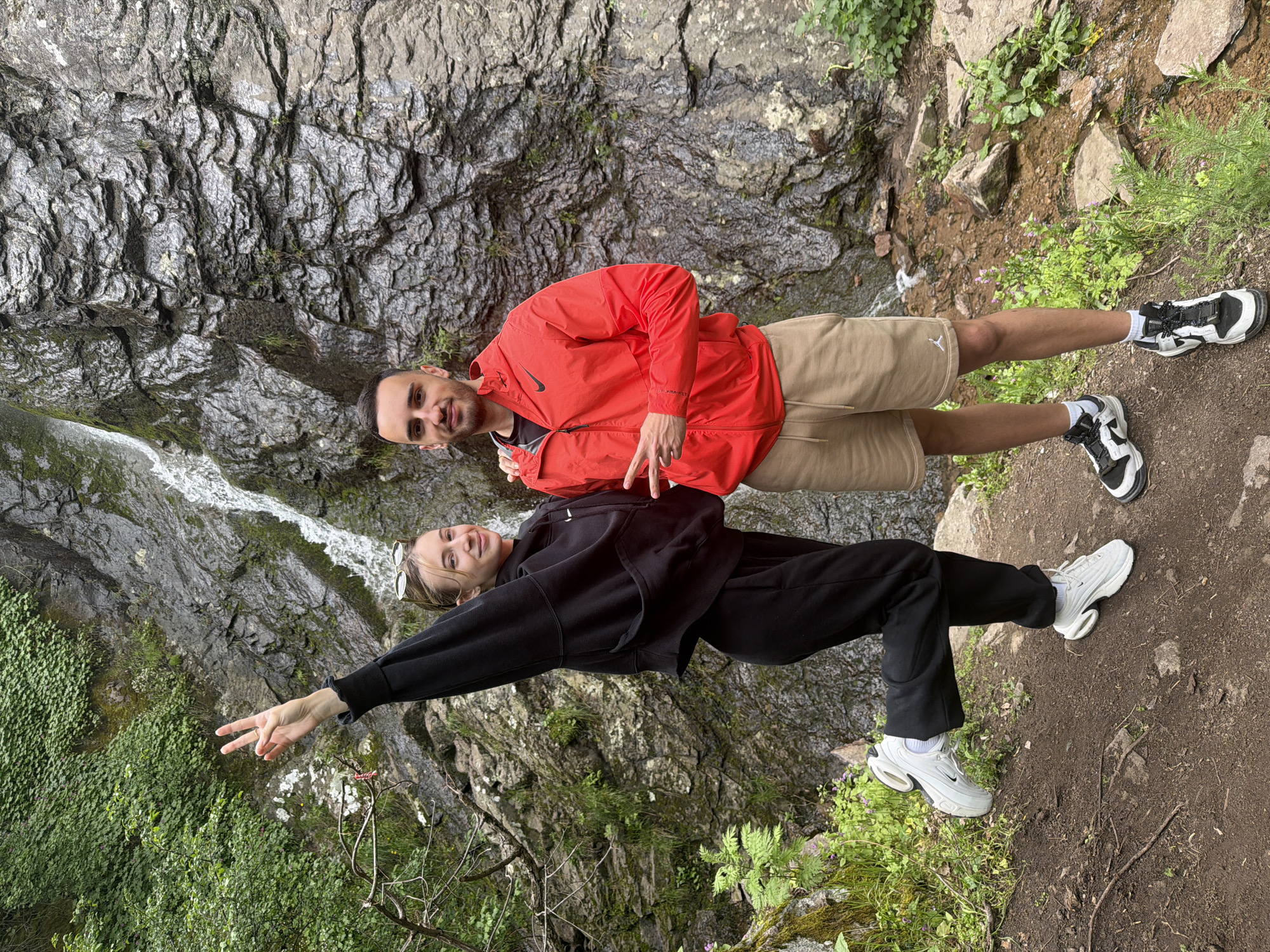 Two people, a male and a female, are positioned in front of a rock face and waterfall near Ustina, Bulgaria. The male is wearing an orange jacket, khaki shorts, and black and white sneakers, making a peace sign. The female is wearing a black sweatsuit and white sneakers, also making a peace sign. They are standing on a dirt path surrounded by green foliage. The rock face and waterfall are in the background.