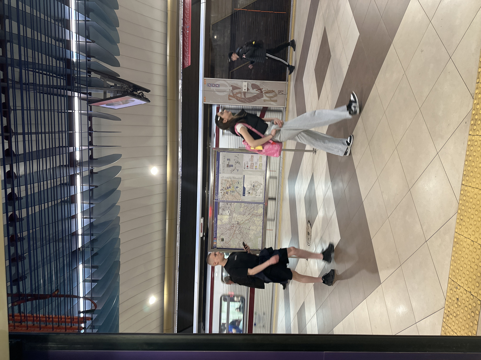The image captures a modern subway station platform, likely in Sofia, Bulgaria, as indicated by visible text on maps and signs. The scene is brightly lit by artificial overhead lighting, suggesting an underground environment or nighttime.
In the foreground, a young man with a shaved head is seen from the side, walking to the right. He wears a black t-shirt, black shorts, and black athletic shoes, and holds a smartphone. Behind him, a young woman with long dark hair is also walking, wearing a black vest over a white top, light grey sweatpants, and black and white sneakers. She carries a pink backpack on her shoulder. Further in the background, near the tracks, another person in a dark jacket with white-striped pants walks away from the camera.
A subway train with red and white stripes is partially visible at the platform edge. The platform floor features a distinctive pattern of large light beige and brown square tiles, laid diagonally. Yellow tactile paving with raised bumps runs along the platform edge.
Along the platform wall, several informational maps are displayed, clearly showing routes and areas of "София" (Sofia) and "Метрополитен" (Metropolitan). Text such as "Градски транспорт София" (Urban Transport Sofia) and "Метролинии" (Metro lines) confirms the location. Above these maps, white panels with abstract grey designs are visible, alongside what appears to be station signage. A digital display screen hangs from the ceiling on the left, which is characterized by an elaborate blue slatted architectural feature. The overall impression is one of a clean, functional, and modern public transport space.