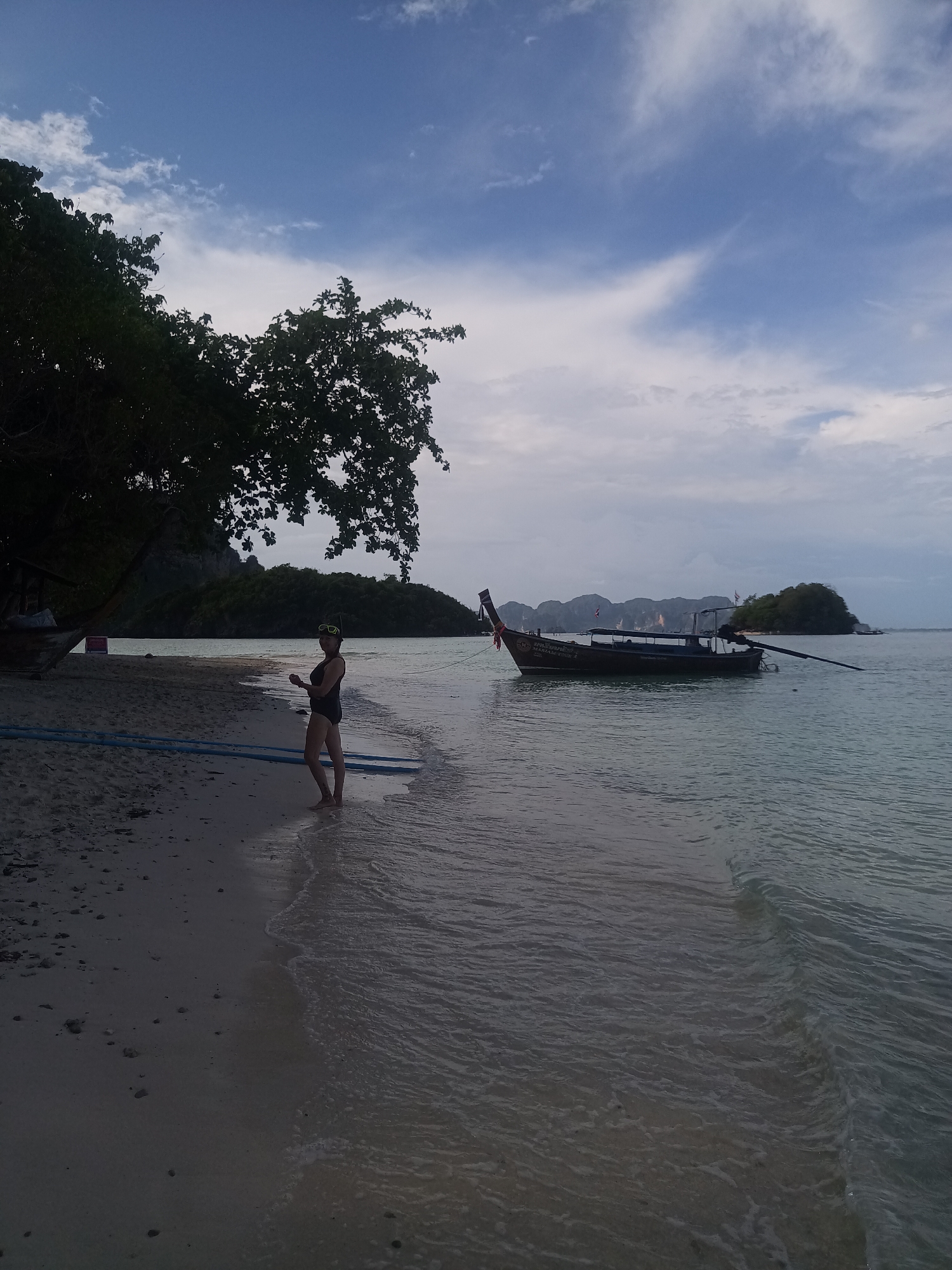 The image captures a beach scene in Ao Nang, Thailand. A woman stands in the shallow water, wearing a black swimsuit and a snorkel mask on her head. She faces towards the water, holding an unidentifiable object in her hand.
A traditional Thai longtail boat is anchored in the water nearby. The boat has Thai flags. The boat has the words "S. SORN SRIN 2" and "MARIAM KNON 2" written on it.
In the background, islands and rocky cliffs are visible. The sky is cloudy, with patches of blue. On the beach to the woman's left, there is a large tree providing shade, and a section of blue pipe rests on the sand. The beach is sandy with some pebbles. The water is relatively calm.