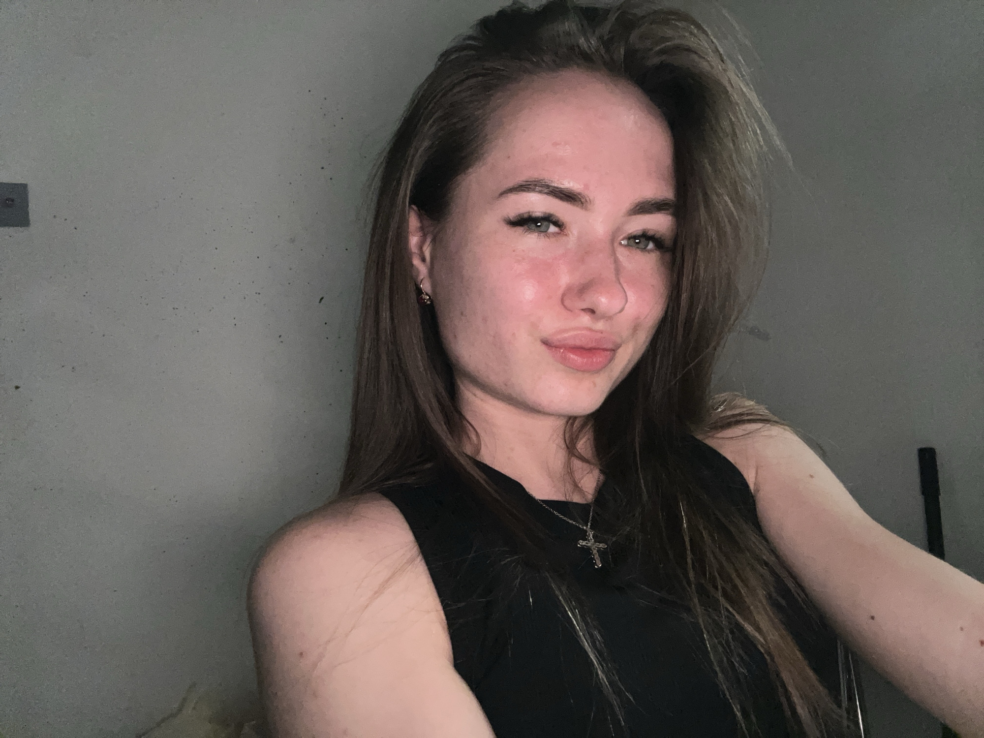 A frontal, medium-close-up shot features a young adult female positioned in an interior setting within the city of Unknown, Unknown. She has long, dark brown hair with lighter highlights, parted on the right side and flowing over her shoulders. Her face is directed towards the camera, displaying a soft, closed-mouth smile. Her light-colored eyes (appearing green/hazel) are open, and her dark eyebrows are well-defined. Her facial skin exhibits areas of redness and minor blemishes, particularly on the cheeks and forehead, along with scattered freckles. A small, dark red stud earring is visible in her left earlobe. She wears a black sleeveless garment with a high neckline and a silver-toned chain necklace with a small crucifix pendant resting on her chest. Her left shoulder and upper arm are visible, while her right arm extends out of frame to the lower left. The background consists of a plain, light gray wall exhibiting a rough, mottled texture with numerous small, dark speckles and a few larger dark marks. In the upper left quadrant of the background, a small, dark square object is affixed to the wall. The lighting appears to be localized, illuminating the subject from the front and slightly above, creating subtle highlights on her forehead, cheeks, and lips. The right side of the background (behind her hair and right shoulder) is darker, possibly indicating a shadow or a deeper section of the room.