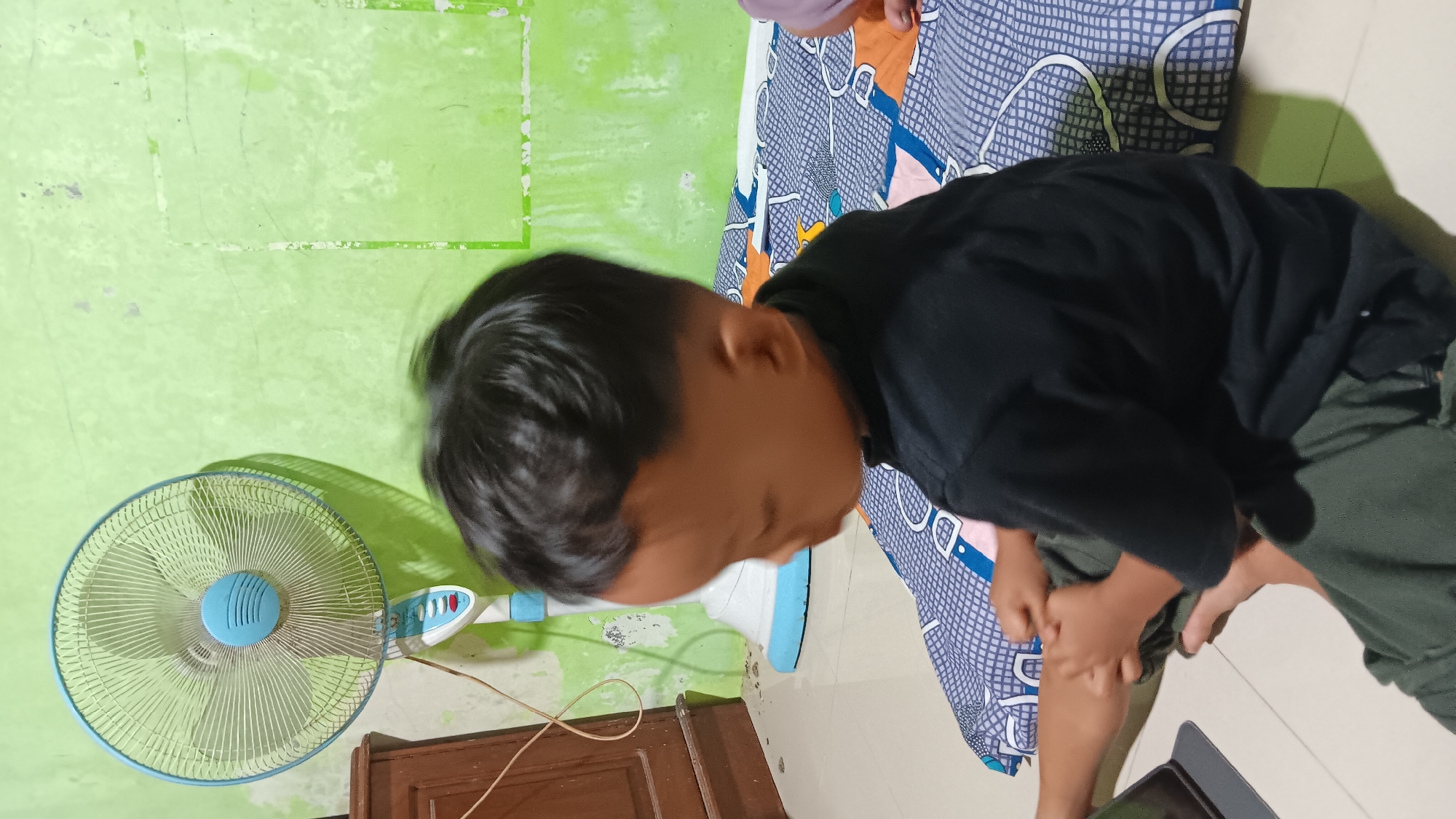 In Sonobekel, Indonesia, a young child, appearing to be a boy, is seated on a patterned mat on the floor. He is wearing a black t-shirt and dark green pants. The child is looking downwards, possibly at a tablet or screen that is partially visible in the lower left corner of the frame. To the left of the child, a wooden cabinet stands against a light-colored wall. In the background, a tall standing fan with a light blue and white body and a blue fan guard is positioned. The walls of the room are painted a vibrant green, with some scuff marks and imperfections visible. The floor is tiled in a neutral color.