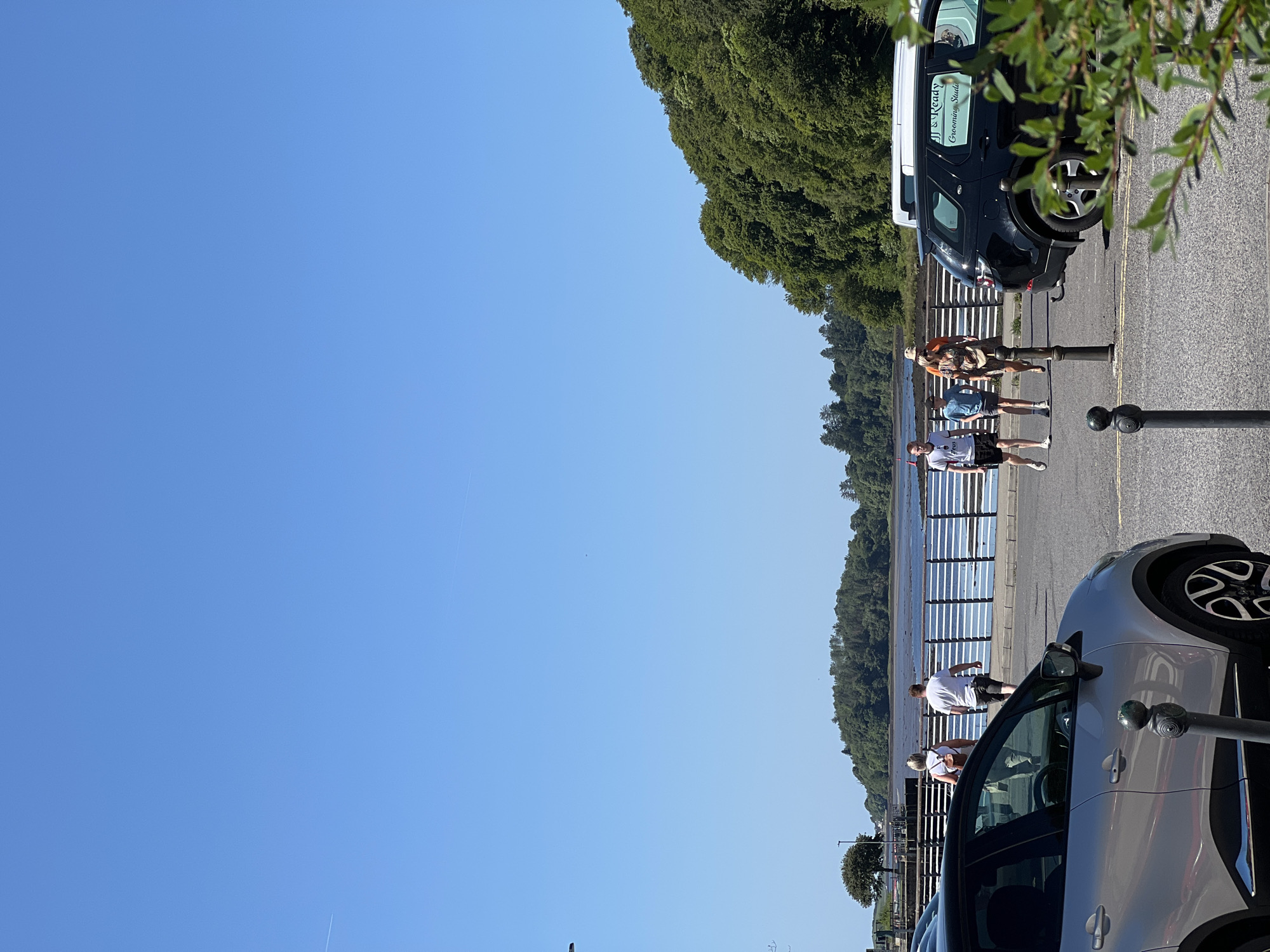 In Donegal, Ireland, under a clear blue sky, a silver car is parked in the foreground to the left. To the right, a black SUV with "JJ & Ready Grooming Studio" written on its side is also parked. Behind the cars, a paved walkway runs parallel to a body of water. A white railing separates the walkway from the water. Several people are walking on the walkway. On the far side of the water, a lush green hillside covered in trees rises up. A lone tree stands on the left side of the image near the water's edge. The overall scene depicts a sunny day in a coastal town.