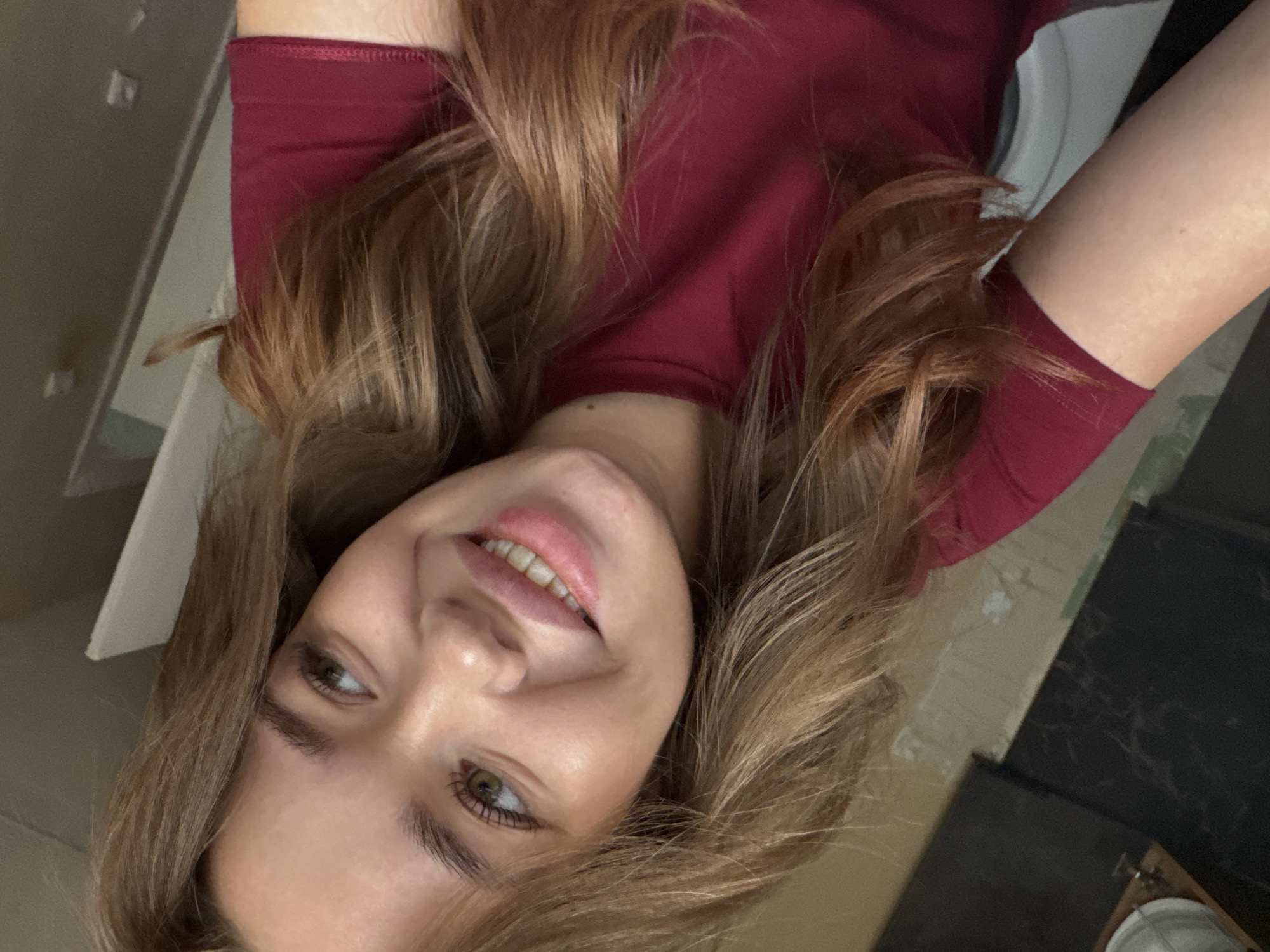 A young woman with light brown hair and green eyes smiles at the camera. She is wearing a maroon t-shirt. The background appears to be an indoor setting with a wall and a doorframe visible. The lighting is soft, creating a warm and inviting atmosphere. The image was taken in Chaikovsky, Russia.