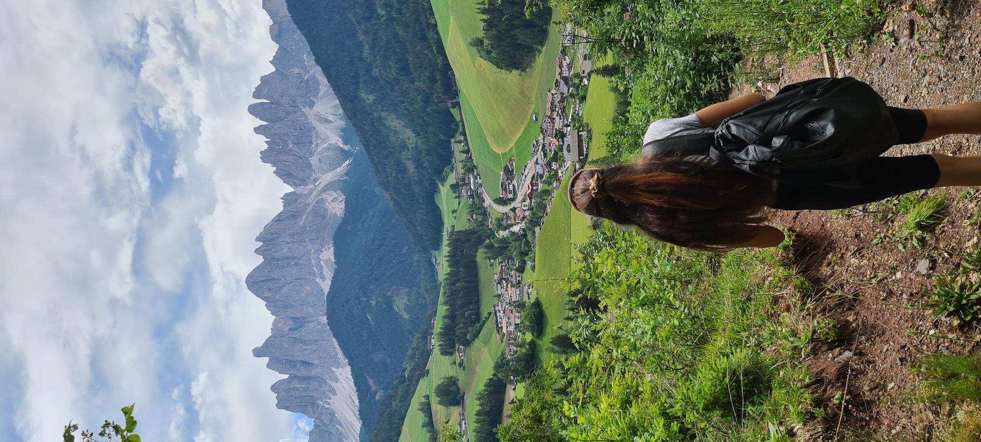 A young woman with long, dark brown hair tied back with a scrunchie, wearing a gray t-shirt and black leggings, walks away from the camera down a dirt path lined with green foliage. She carries a black backpack. In the background, a picturesque village with red-roofed buildings is nestled in a valley, surrounded by rolling green hills and dark green forests. Majestic, jagged mountain peaks with rocky slopes and some patches of snow dominate the skyline under a cloudy sky. This scene is characteristic of the Funes Valley in South Tyrol, Italy, specifically the area near the church of St. Magdalena in Santa Maddalena, known for its stunning Dolomite vistas.