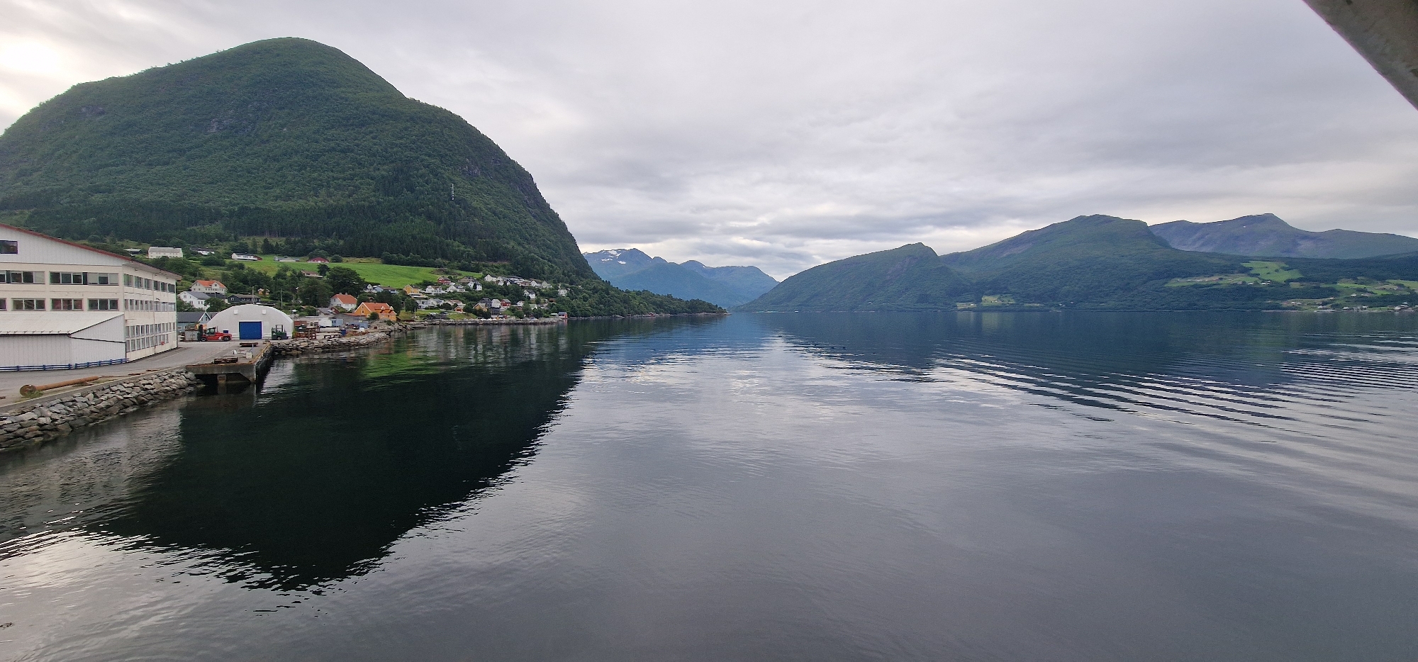 The image depicts a serene waterscape with a village nestled against lush green mountains, likely in Volda, Norway. In the foreground, a calm body of water reflects the overcast sky and the surrounding landscape. On the left side of the image, a prominent building complex with a white facade and numerous windows is situated along the waterfront. Adjacent to this building is a dock with a white arched structure, and a few small vehicles are visible. The buildings of the village, characterized by colorful roofs, are scattered along the base of a steep, tree-covered mountain. Further in the distance, other mountains rise, some with snow-capped peaks, under a cloudy sky. The overall scene conveys a sense of tranquility and natural beauty.