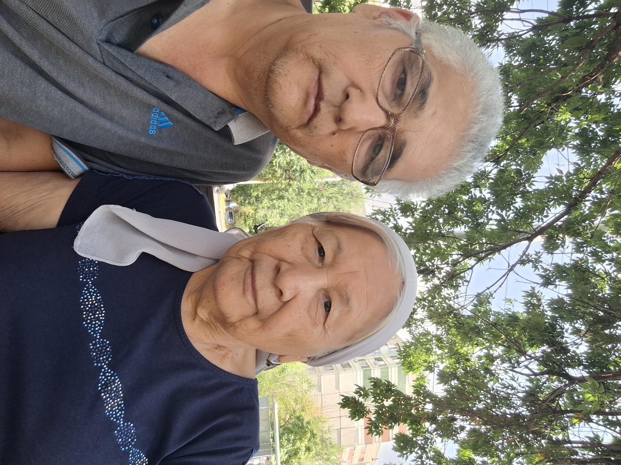 This is a selfie of an elderly Kazakh couple sitting outdoors under a tree with green leaves. The man, on the left, wears glasses and a grey polo shirt with a blue Adidas logo. The woman, on the right, wears a navy blue shirt with a decorative beaded pattern and a light grey headscarf. In the background, there are trees, a building, and a clear sky. The scene appears to be in a park or residential area.