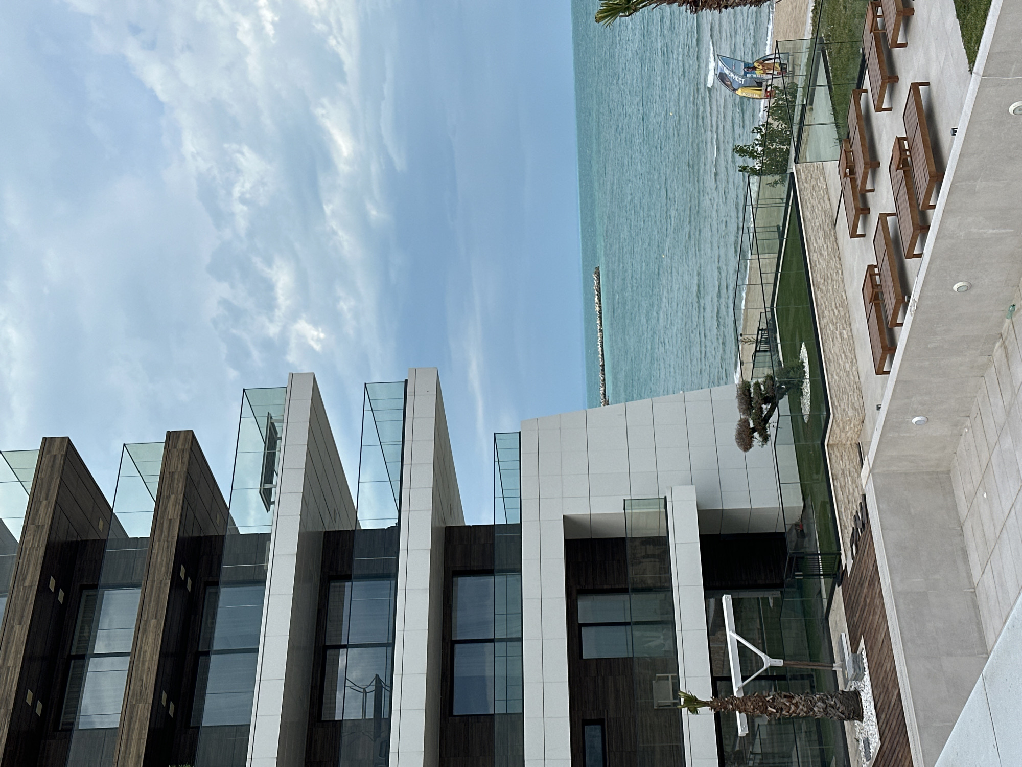 The image depicts a modern architectural building situated in Varna, Bulgaria, near the sea. The building features a facade with dark wood panels, white vertical columns, and glass elements that extend upwards, creating a stepped design. Large windows are set into the building's structure.
The sea is visible in the background, with its blue water extending towards the horizon under a cloudy sky. A portion of a patio or terrace is visible on the right side of the building, complete with lounge chairs and landscaping. A palm tree is positioned near the building's edge.
There are no people visible in the image. The overall impression is of a high-end, modern development in a coastal location.