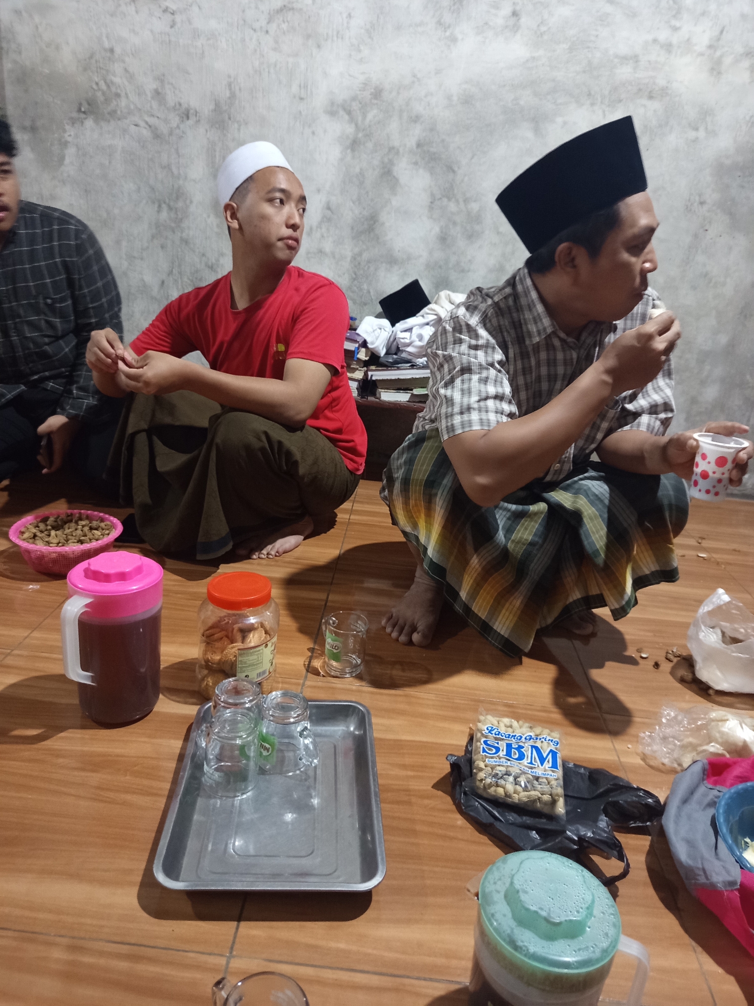 The image depicts a group of men, likely Muslim, sitting on a wooden floor in what appears to be a humble indoor setting, possibly in Dermo, Indonesia. One man on the left wears a white peci (cap) and a red t-shirt, while another man in the center, also wearing a black peci, is dressed in a plaid shirt. Both men are in a squatting position. In the foreground, there are several items on the floor, including a pink-lidded pitcher filled with a dark liquid, a clear jar with cookies, and a metal tray holding three small glasses. A plastic bag containing peanuts and a light blue pitcher are also visible. The background shows a textured wall. The overall atmosphere suggests a casual gathering, possibly a meal or a moment of rest.